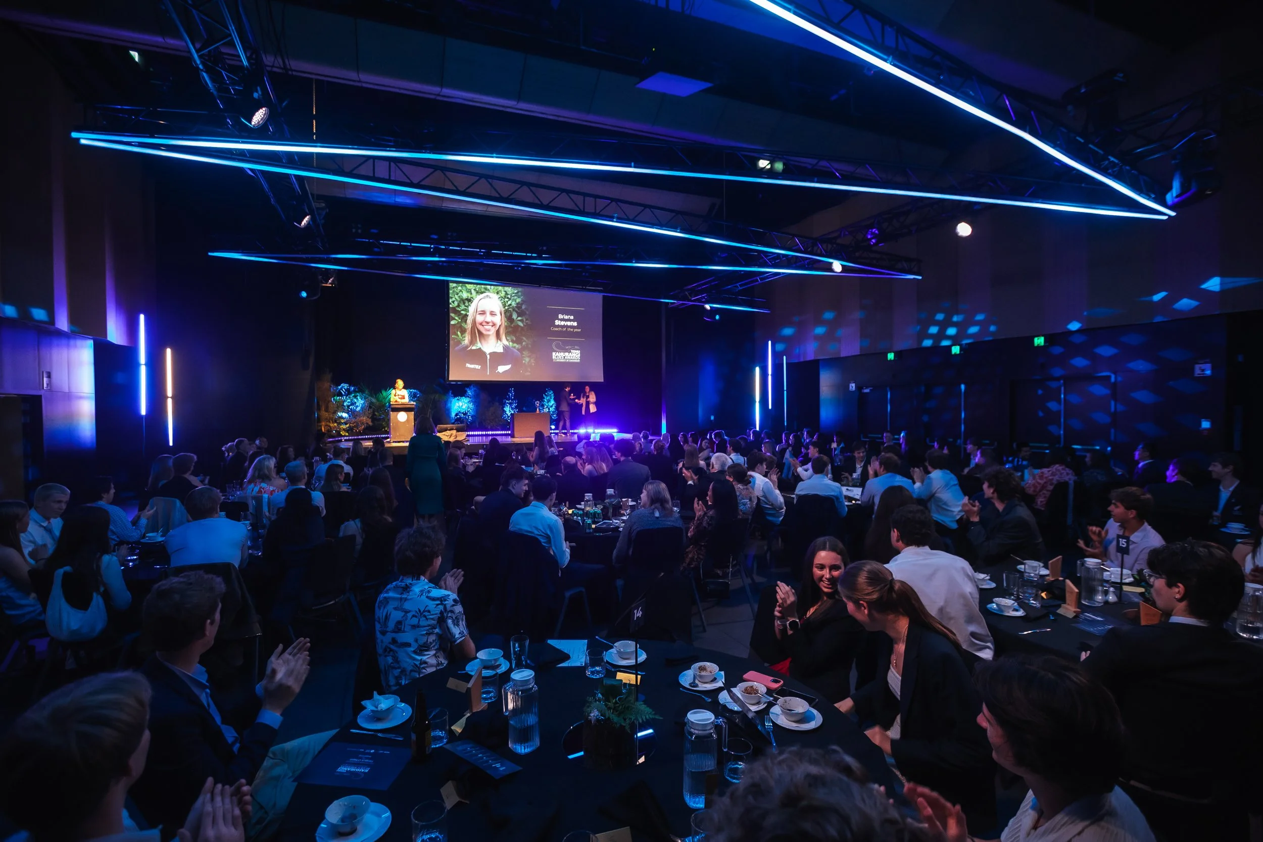 A large dark conference room filled with many people seated at round table. The room is illuminated with blue and purple lighting from neon-like accents and stage lighting. On stage, there is a large screen displaying a smiling woman named Briana Stevens, listed as a coach of the year, with an image of greenery behind her. The stage has a podium, and there are people standing on stage. The audience appears to be clapping.