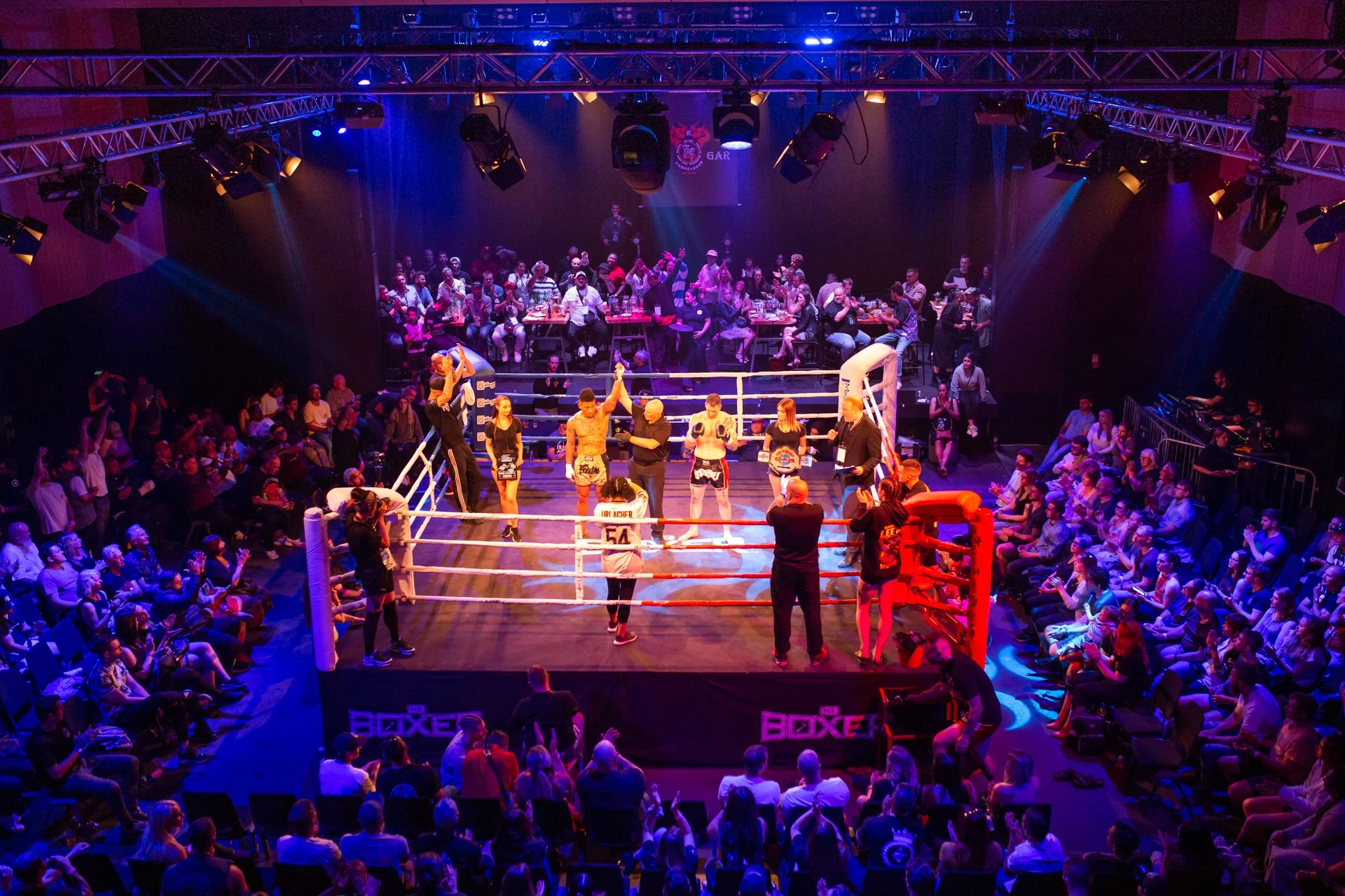 A boxing match taking place in an arena filled with spectators. The fighters are in the ring with a referee and corner team members. Audience members are seated around the ring and in tiered seating, with some standing and cheering. The arena is illuminated with colorful lights, and the event has an energetic atmosphere.
