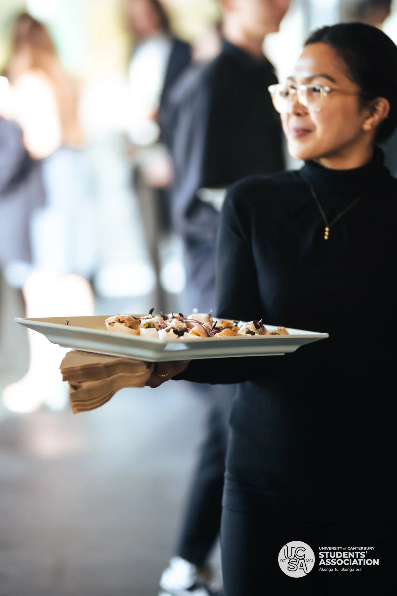 A person wearing glasses and a black top holds a tray with appetizers, likely at a social event. Blurred people are seen in the background, suggesting a gathering or reception.