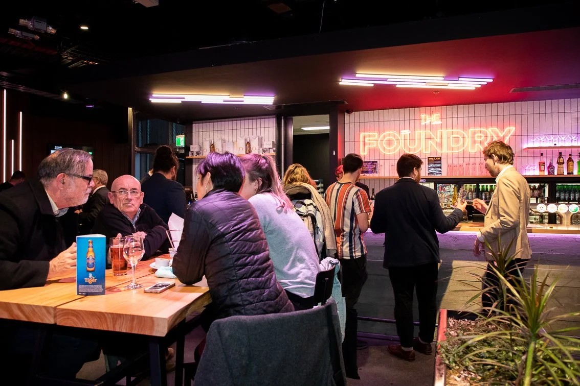 People sitting at a restaurant bar with a neon sign that reads 'The Foundry' in the background.