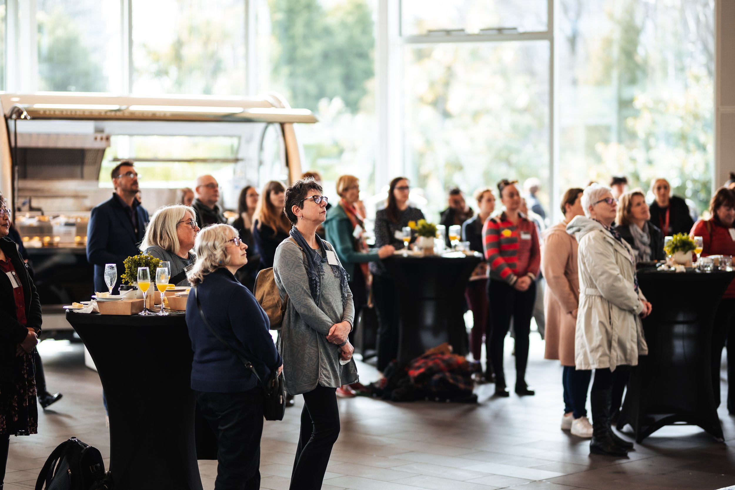 People attending a conference or presentation in a well-lit room with large windows, some standing and some sitting at tables with flowers and drinks.