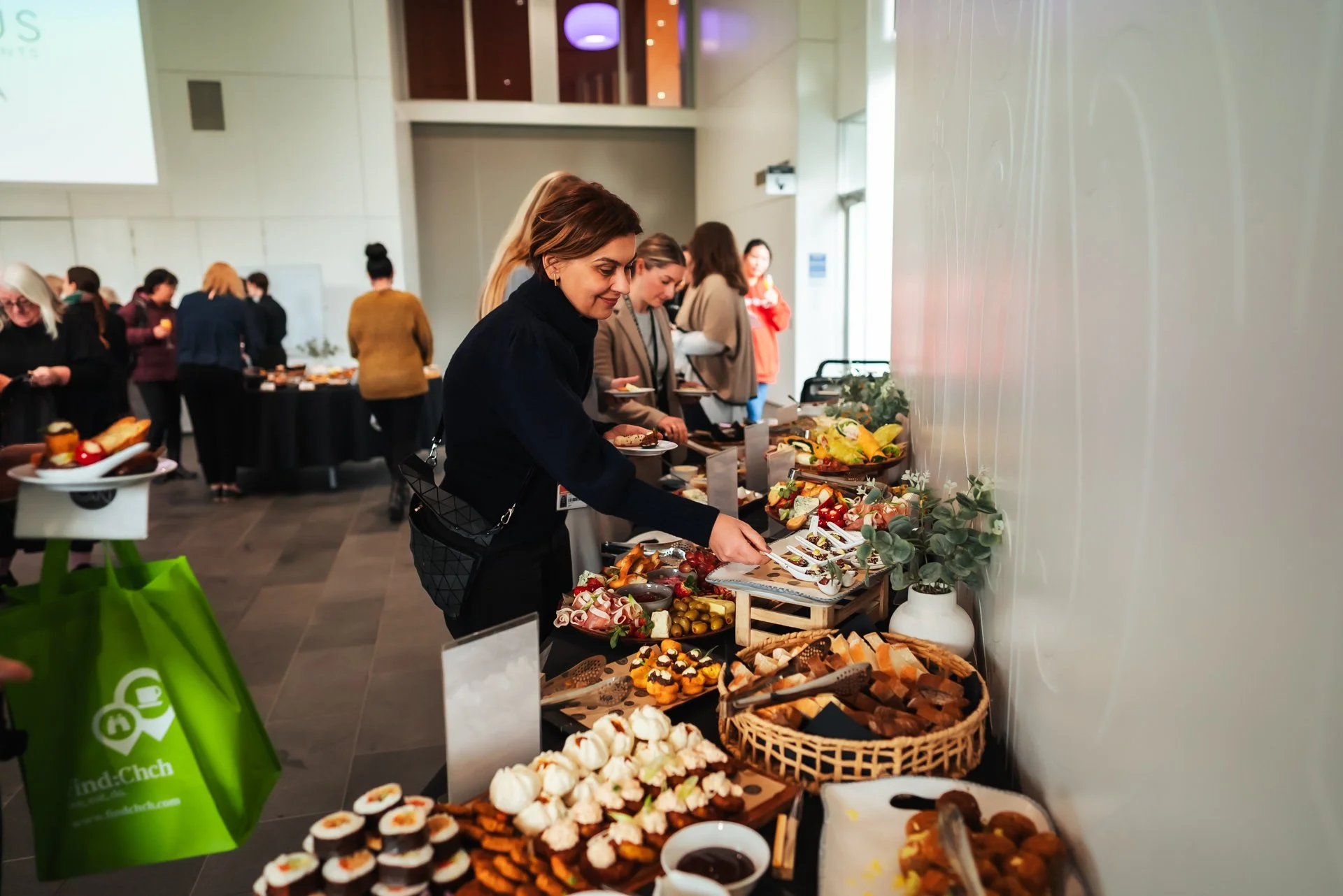 Woman selecting food from a buffet table at an indoor event, with various appetizers and desserts on display and other guests in the background.