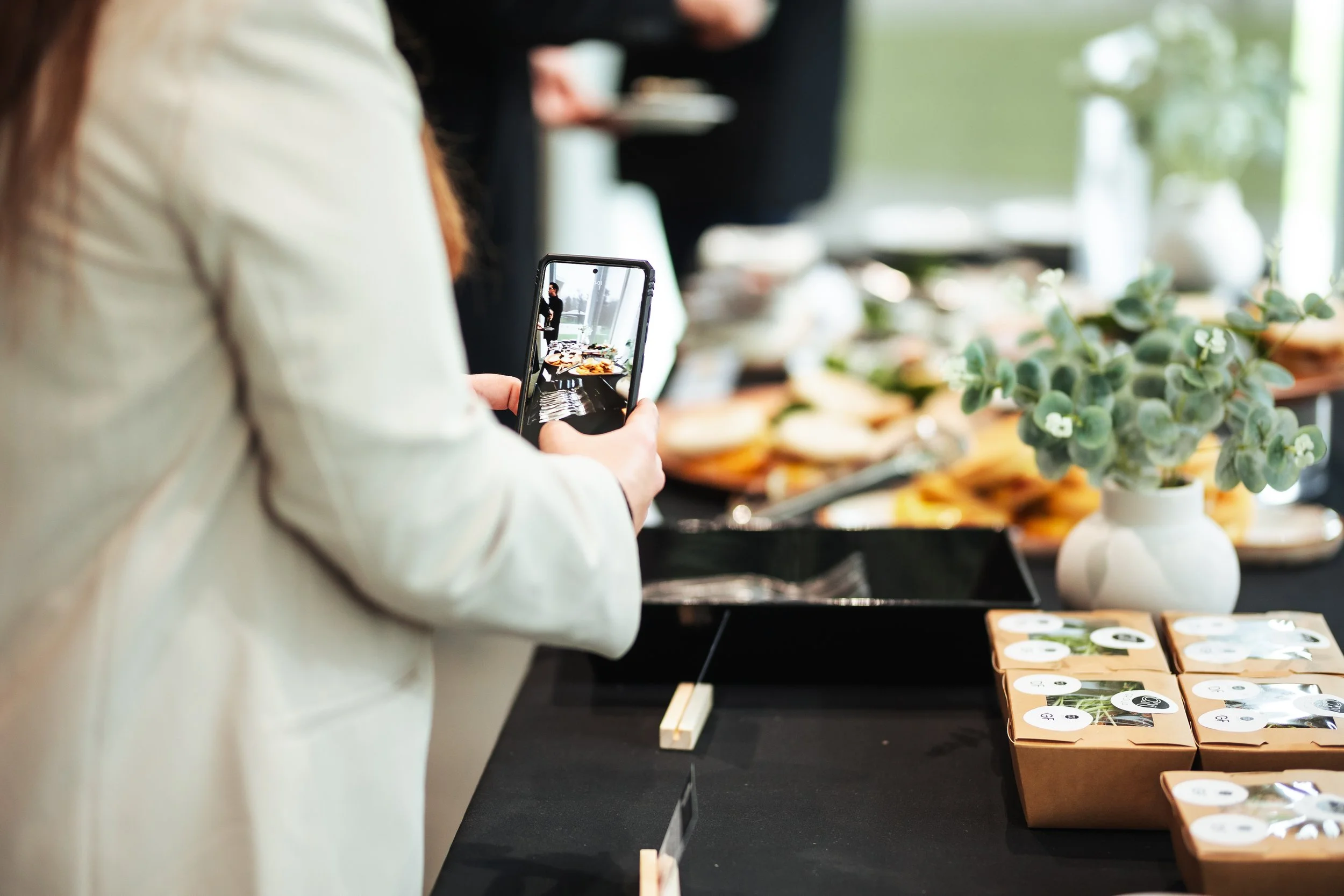 Person taking a photo of items on display table at a market or store with a smartphone, with trays of food and small potted plants visible on the table.