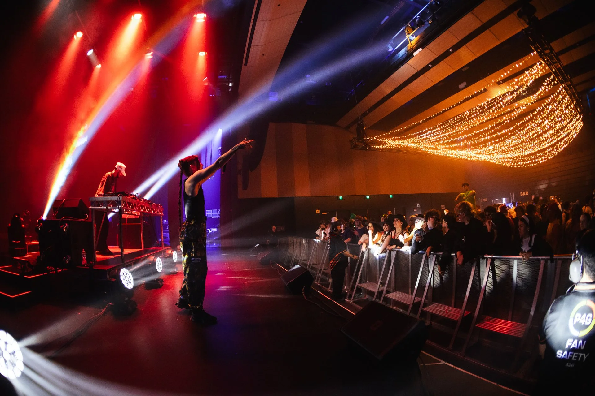 Concert scene with colorful stage lights, a DJ on a platform, a performer with braided hair pointing towards the audience, and a crowd of fans behind a barrier, with decorative string lights on the ceiling.