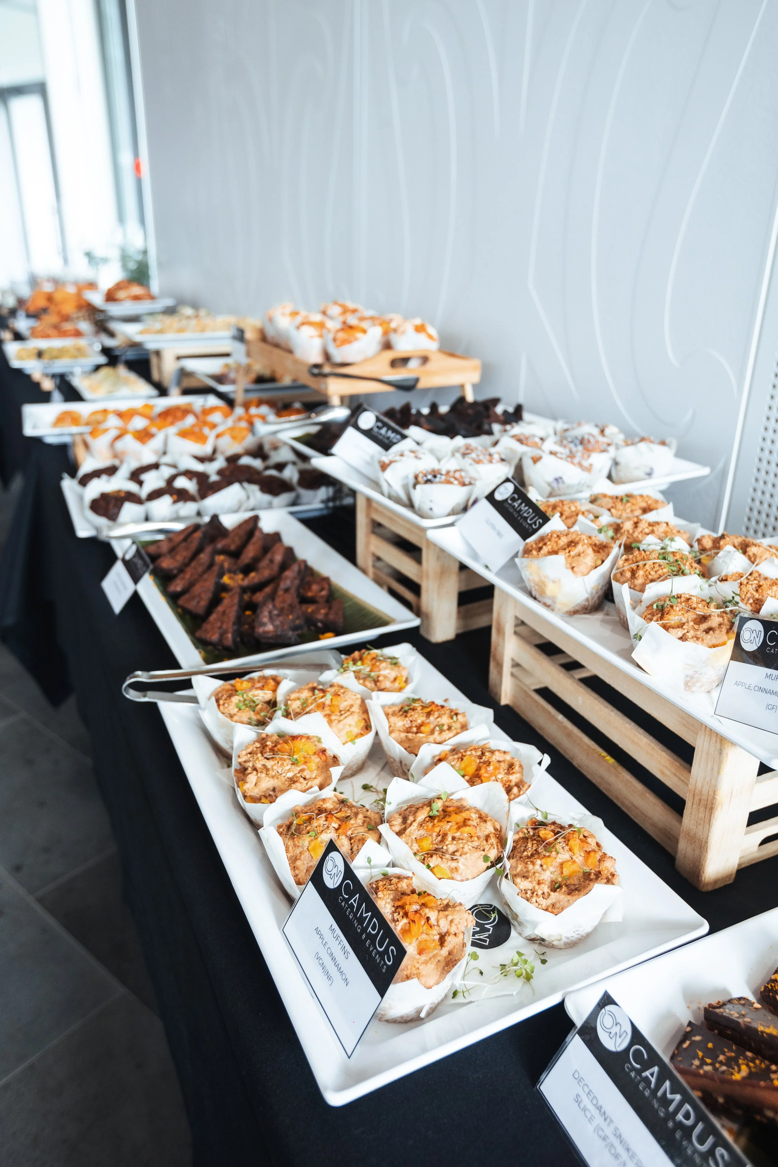 A buffet table with various baked goods and snacks, including muffins, cookies, and sliced items, labeled with tags and displayed on trays and wooden stands.