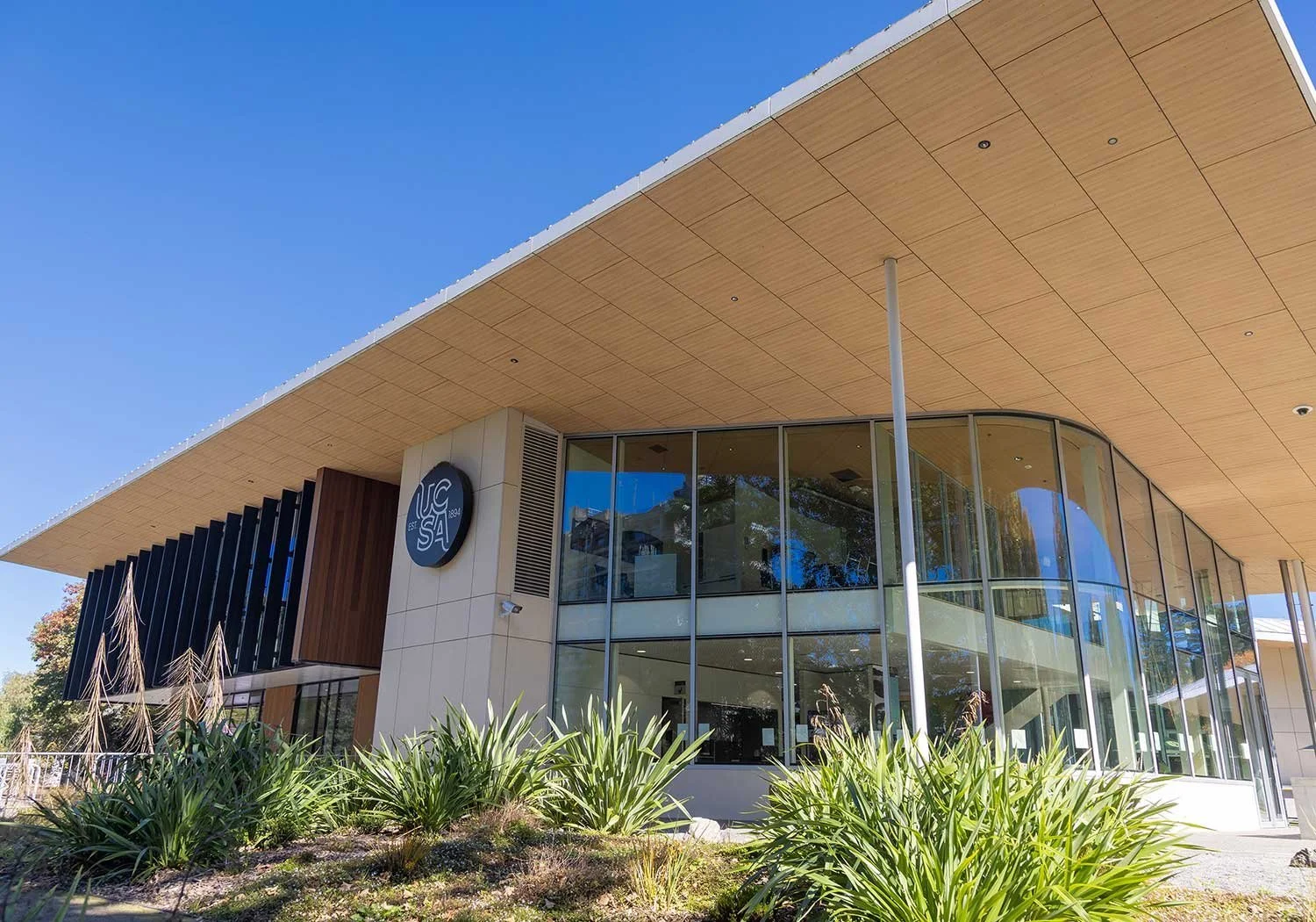 Exterior of a modern building with large glass windows, wooden accents, and a circular sign with the UCSA logo, surrounded by greenery and clear blue sky.
