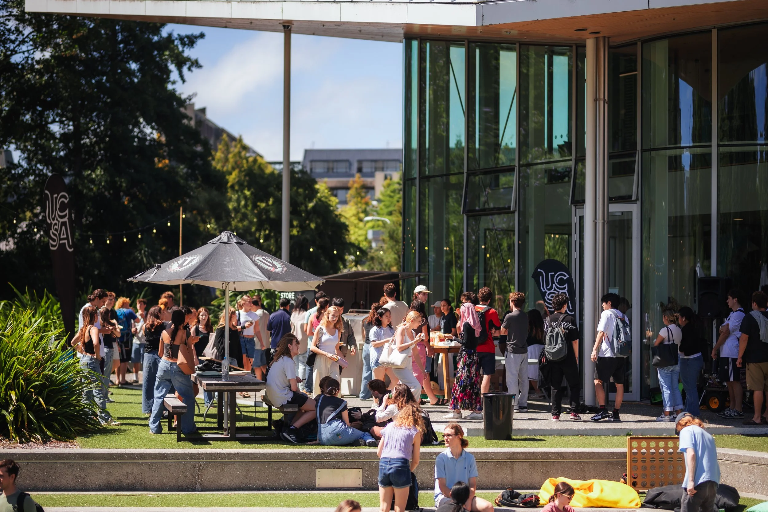 A large group of people gathered outdoors, many standing and some sitting near tables, in front of a modern glass building with trees and a blue sky in the background.