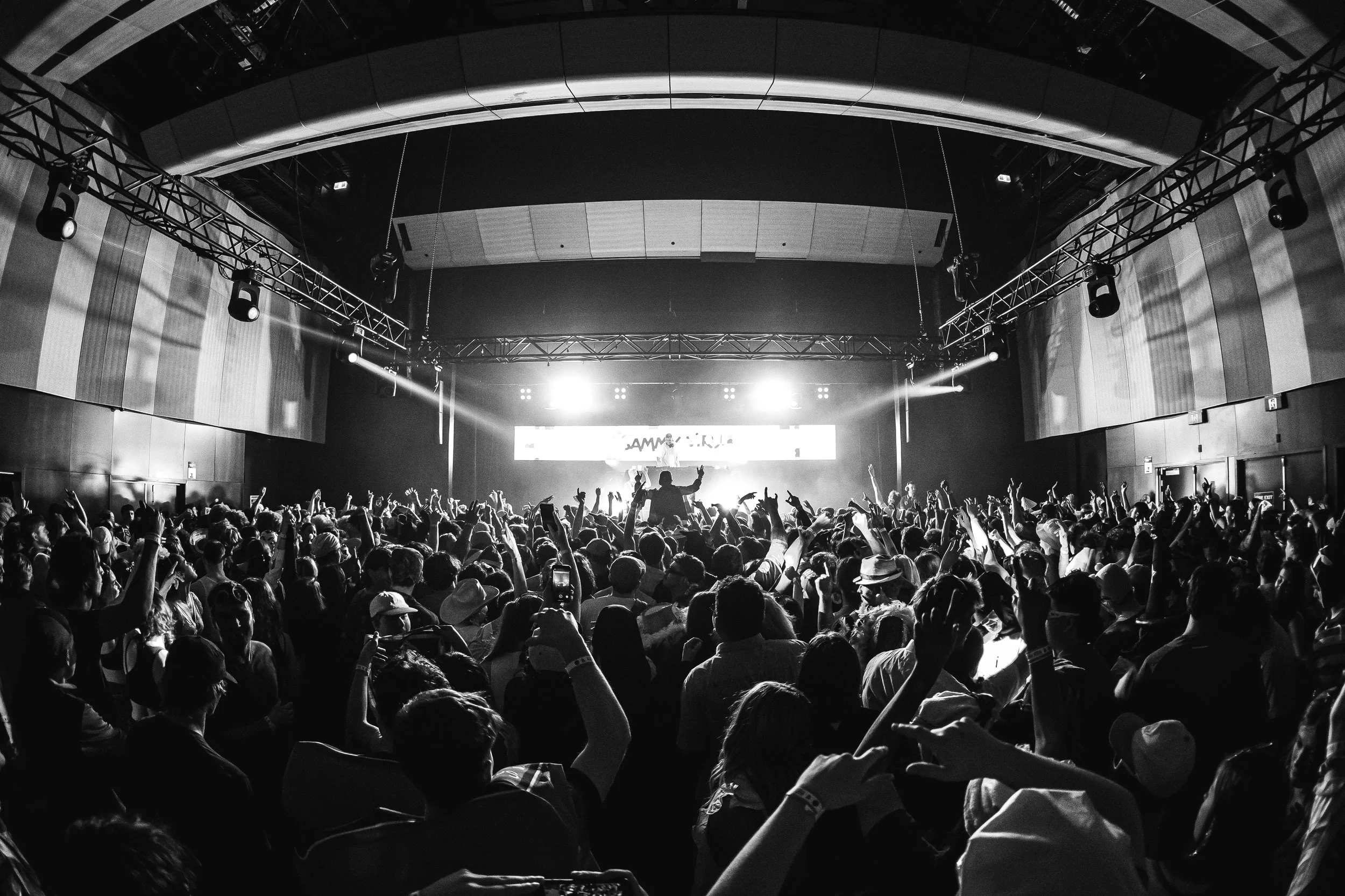 A large crowd of people at a concert or event in a spacious indoor venue with stage lighting and a performer on stage.
