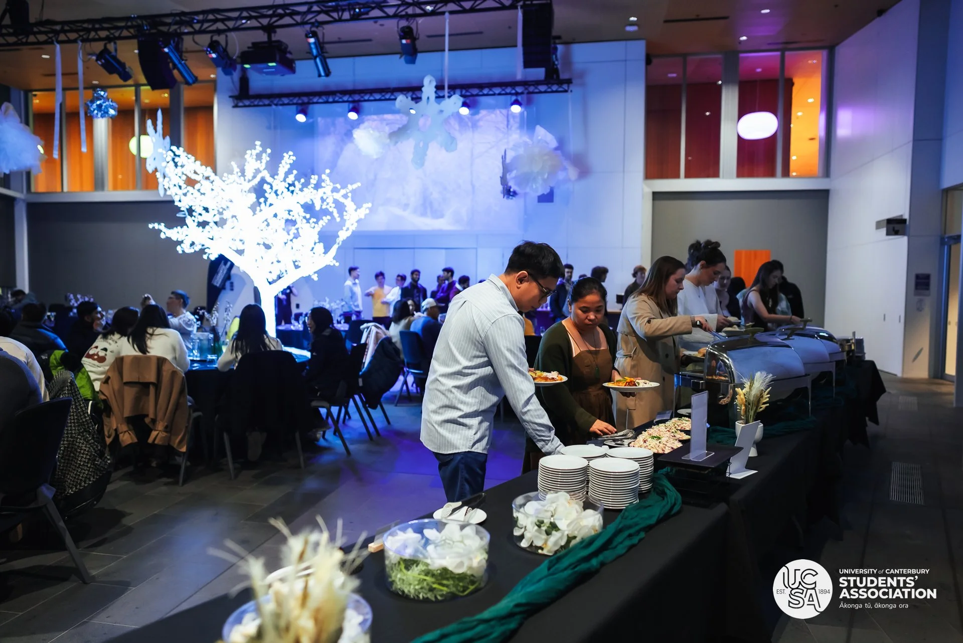 Students at a university event serving food at a buffet table with people in line and seated guests in a large, decorated hall with lighting effects and a projection on the wall. Logo of the University of Canterbury Students' Association in the corner.