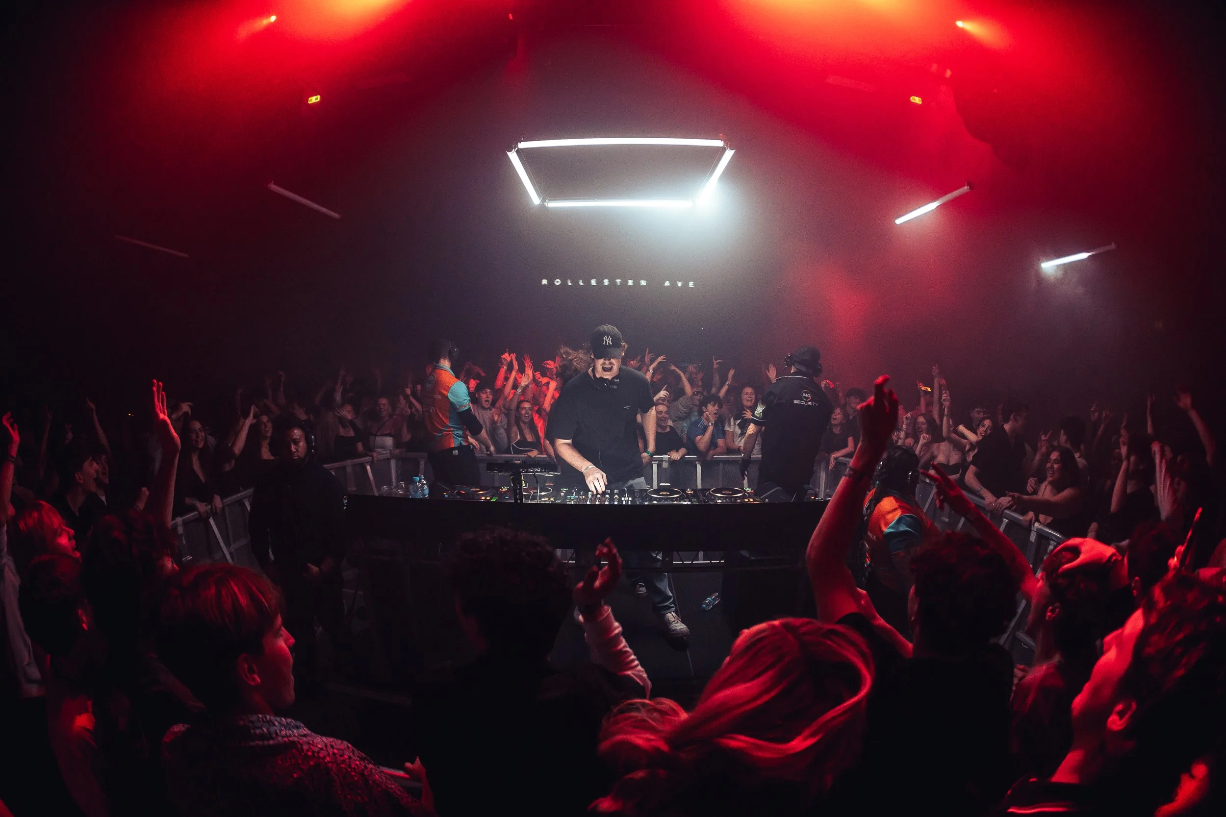 A DJ is performing on stage at a nightclub with a crowd dancing and raising hands. The stage has a bright neon frame and the words 'ROLESSTER AVE' displayed above the DJ.