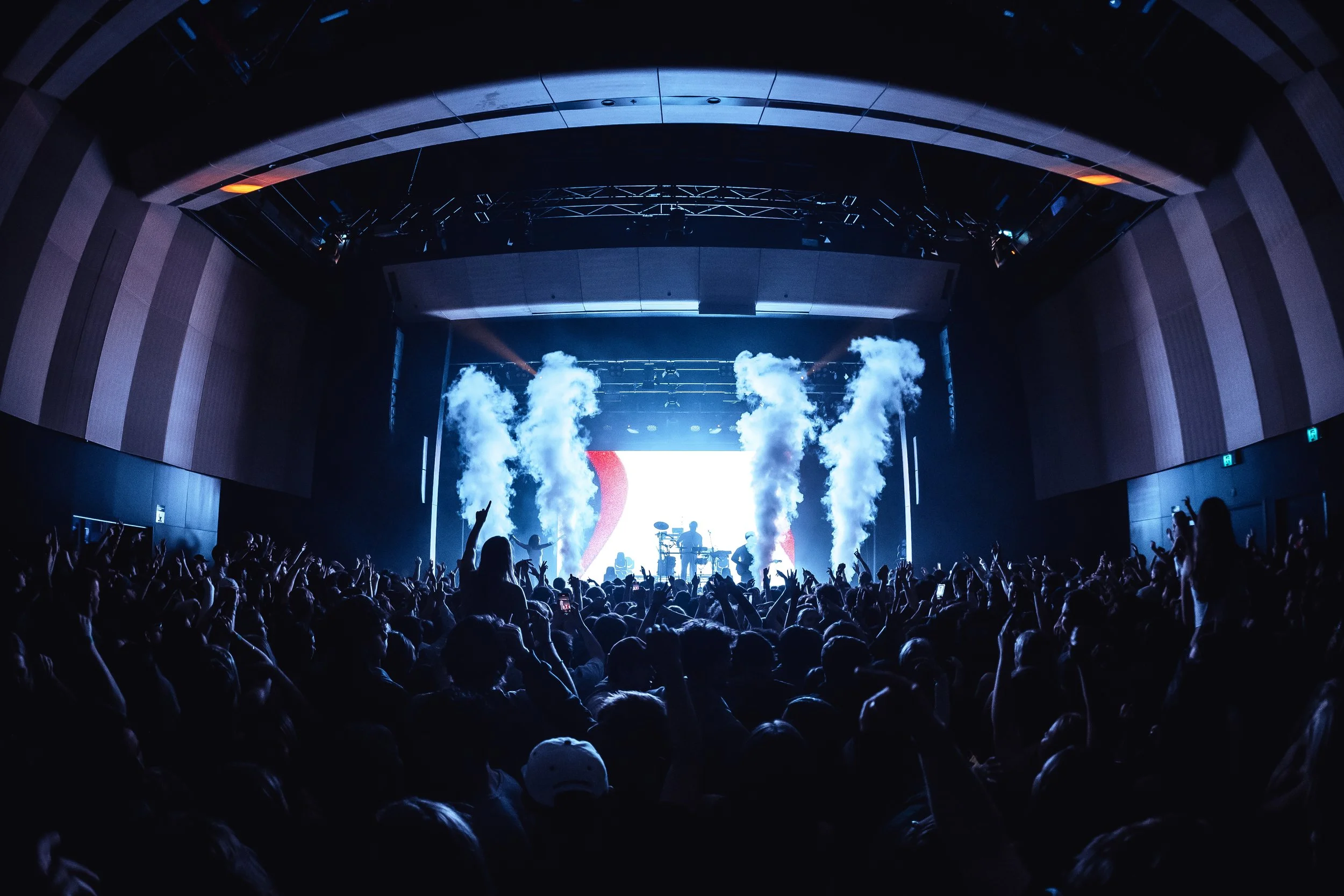 A concert stage with performers, smoke effects, and audience raising their hands in a dark indoor arena.