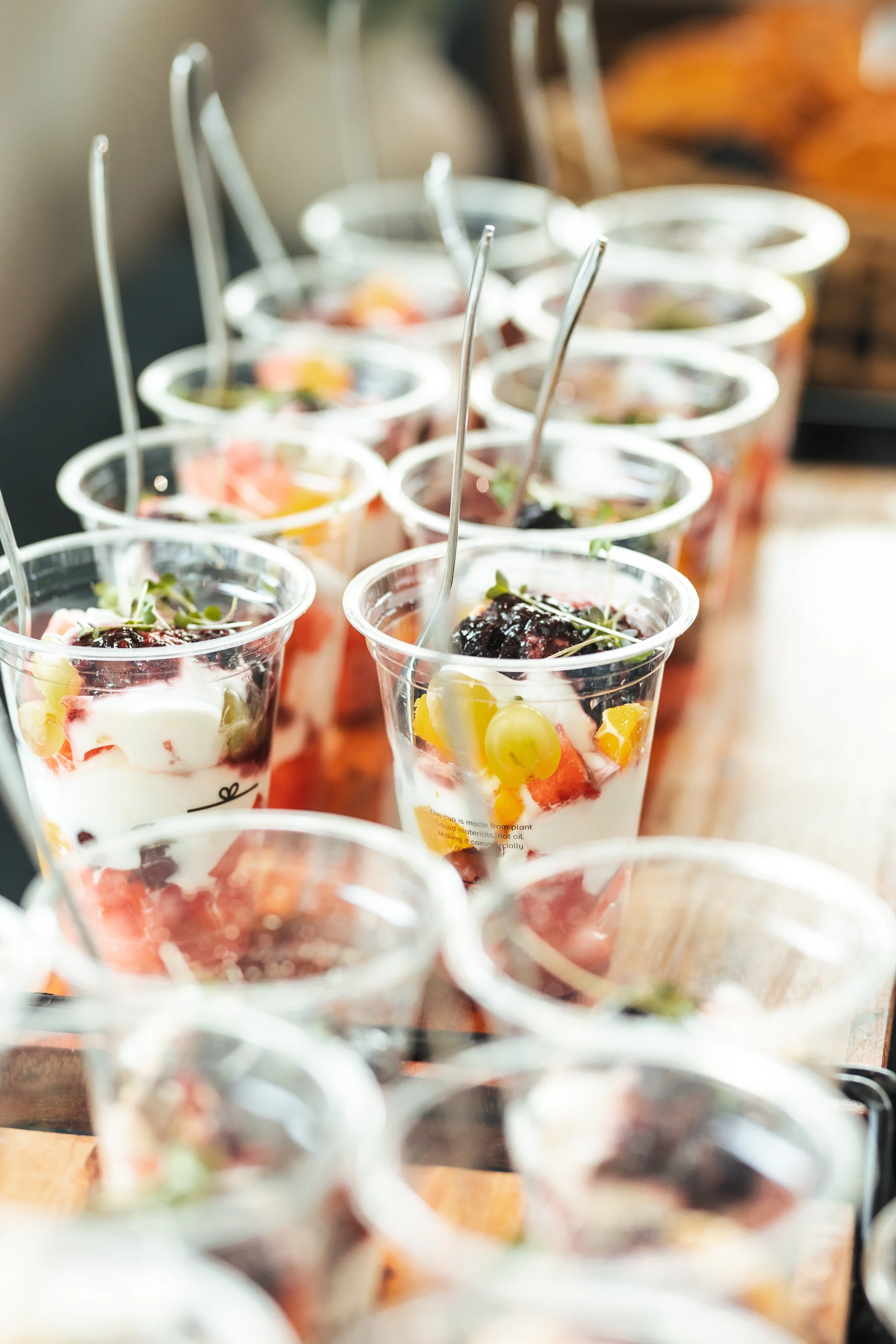 Several small clear plastic cups filled with fruit salad, each with a small spoon, arranged on a wooden surface.