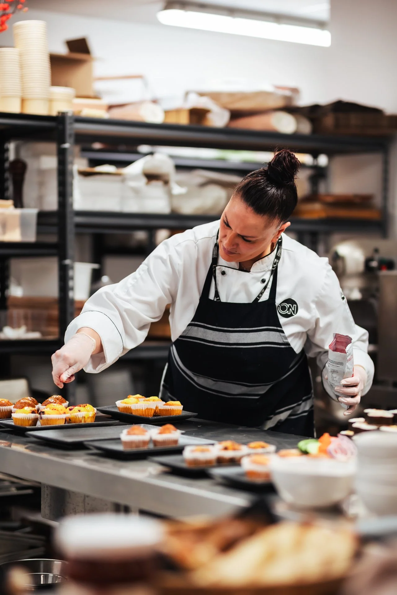 A chef in a white coat and black apron preparing desserts in a commercial kitchen, with trays of cupcakes and pastries on a stainless steel counter.