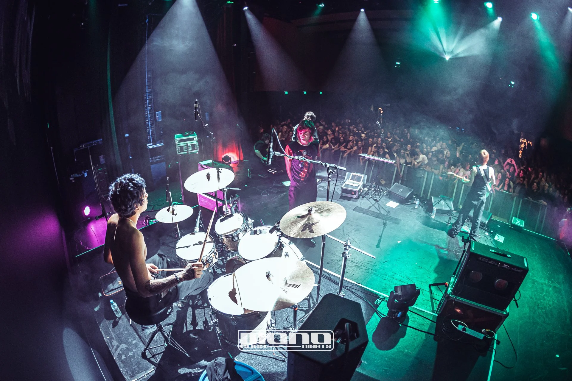 A live music performance on stage with a drummer, guitarist, and singer, facing an audience in a darkened venue with colorful stage lighting.