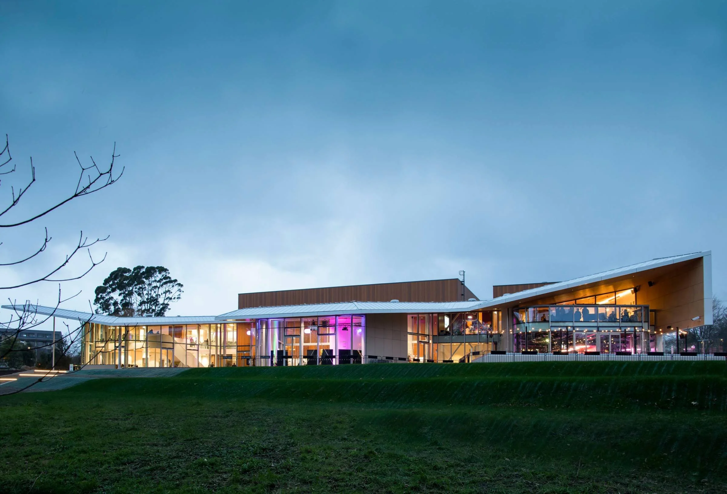 A modern building with large glass windows illuminated from inside, set against a darkening sky with trees in the background and a grassy area in the foreground.