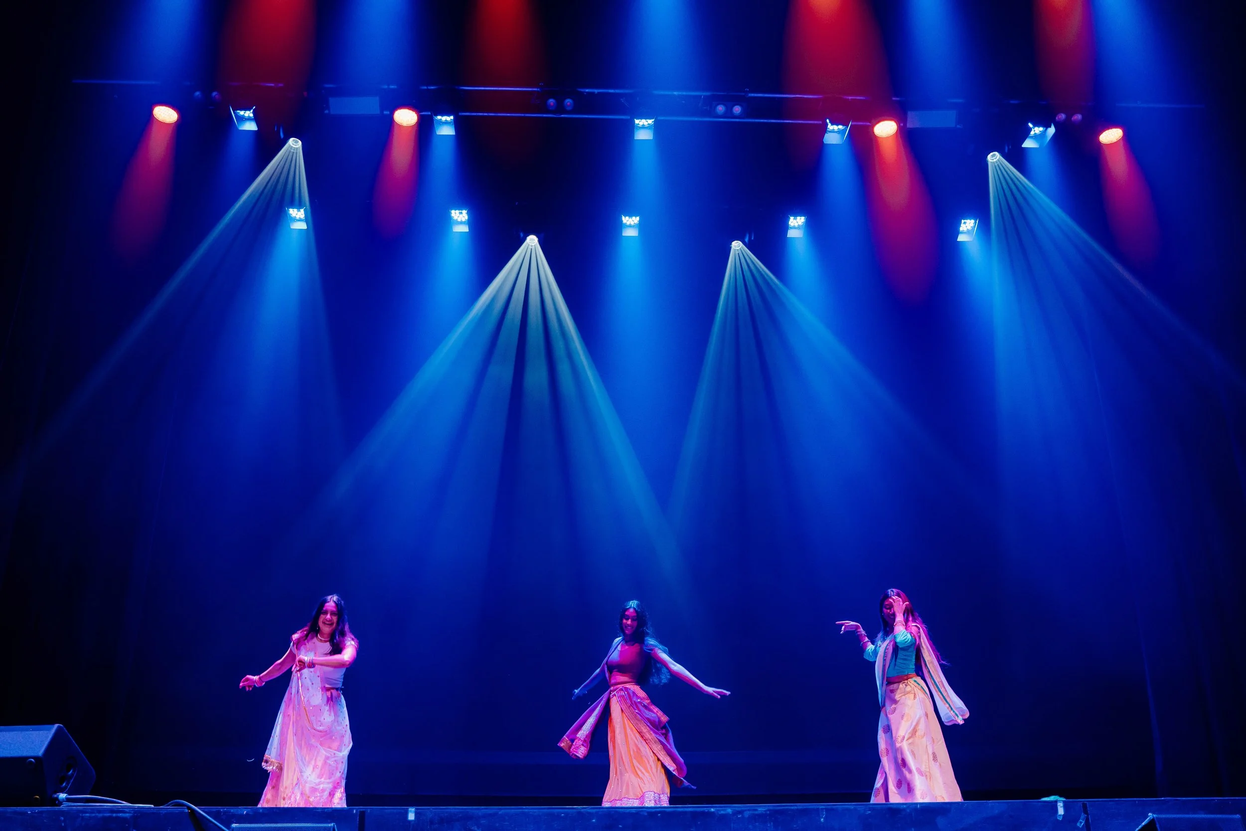 Three women performing a dance on stage with colorful lighting, wearing traditional dresses.