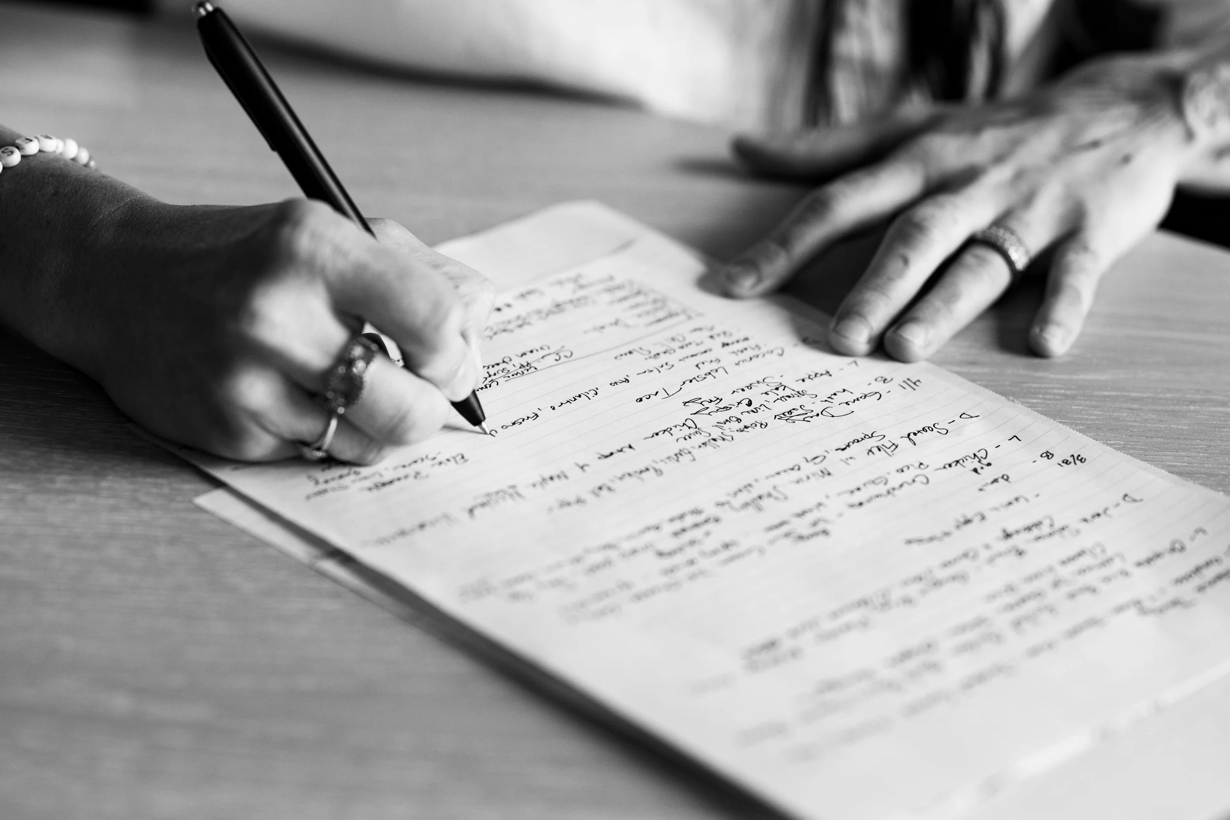 Black and white photo of a person writing handwritten notes in a notebook on a wooden table, with a blurred object in the background.