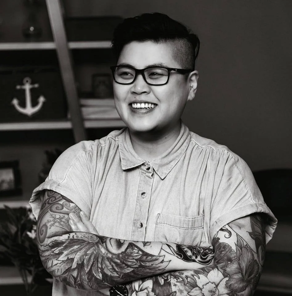 Black and white portrait of a person with short hair, glasses, and tattoos on their arms, smiling with arms crossed, indoors with shelves and an anchor decor in the background.