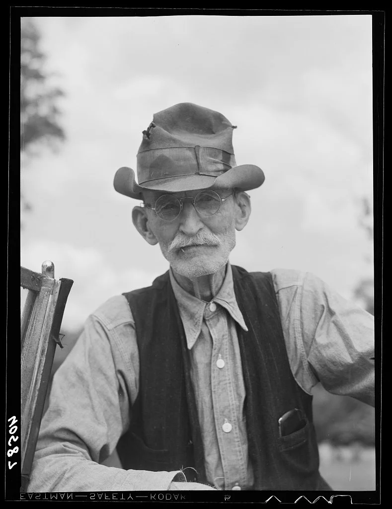 Black and white photo of an elderly man with a white mustache and beard, wearing round glasses, a weathered cowboy hat, a button-up shirt, and a vest, outdoors with cloudy sky and trees in the background.