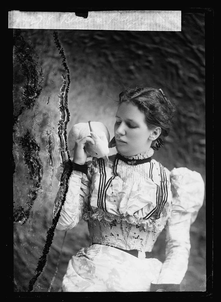 A woman in vintage clothing, with a high-collared blouse and ornate details, holding an apple and a pen as she looks at it thoughtfully.