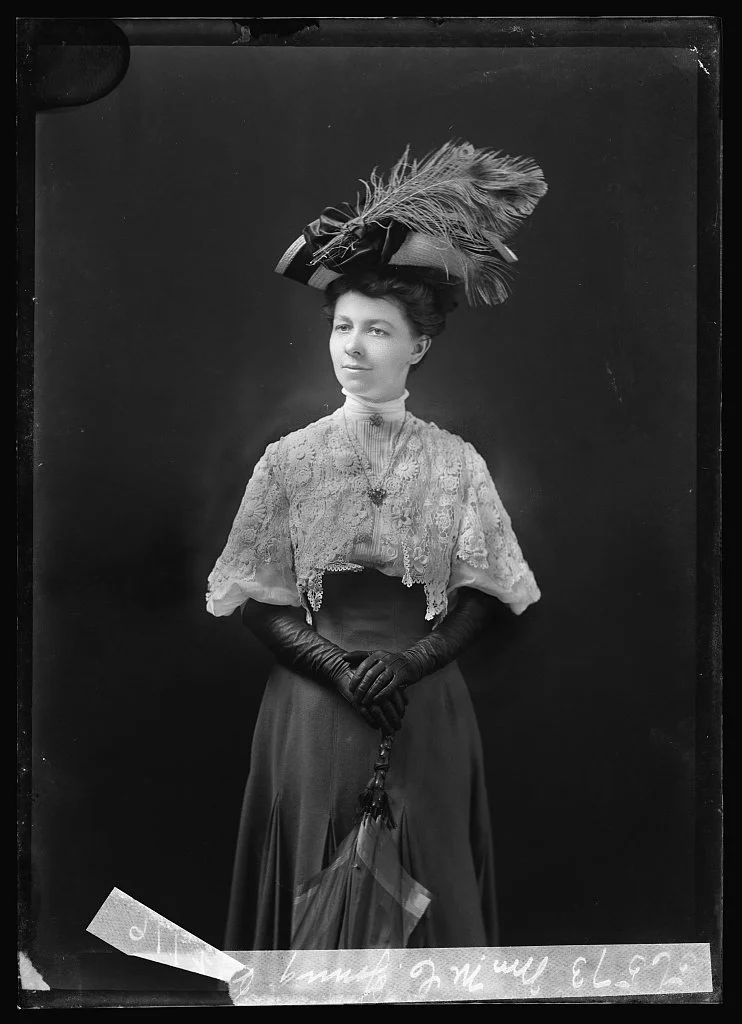 A black and white photograph of a woman in vintage clothing, wearing a large feathered hat, lace top, long skirt, gloves, and holding a parasol.