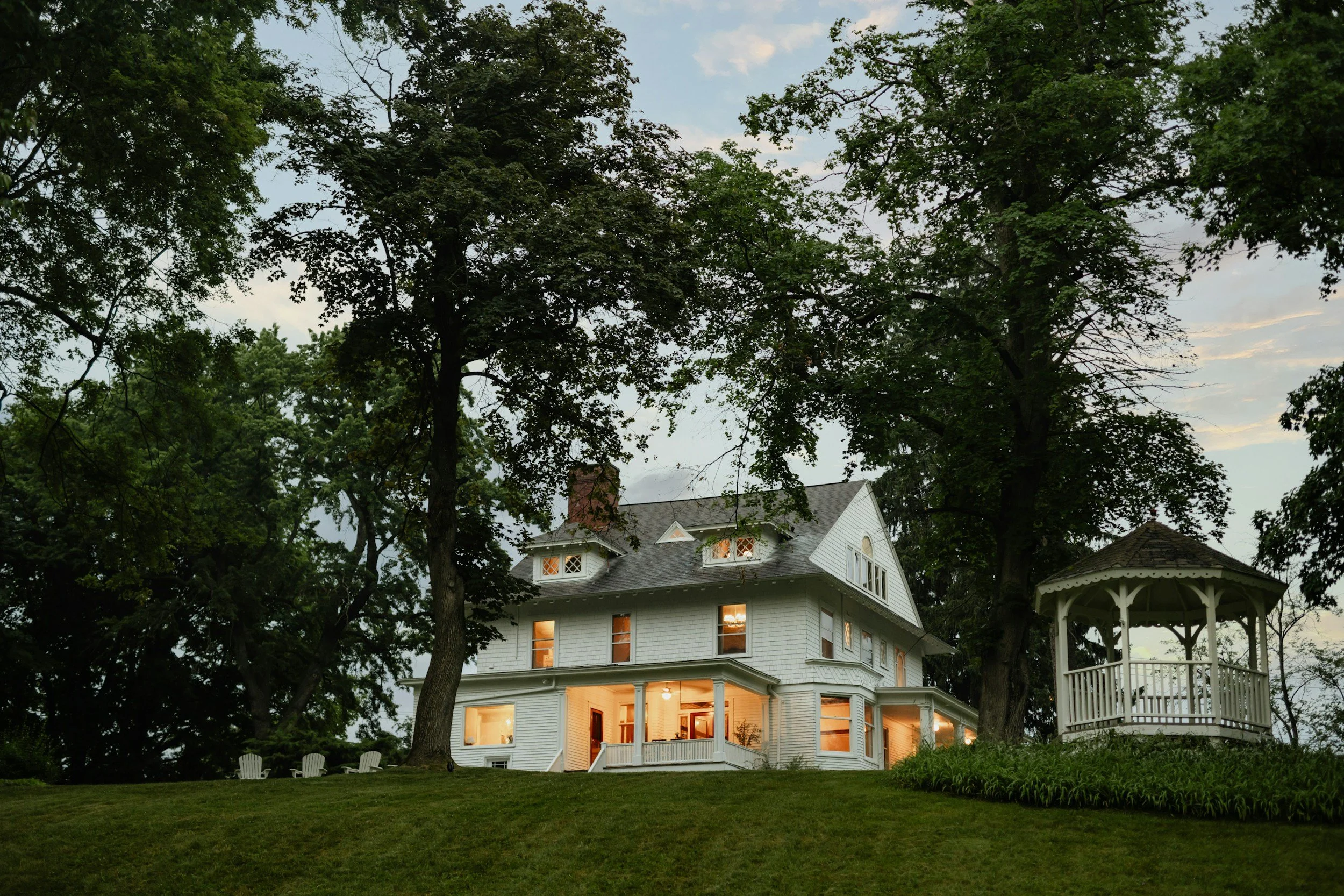 A large white house with multiple stories and illuminated windows, surrounded by tall trees in a lush green yard during dusk.