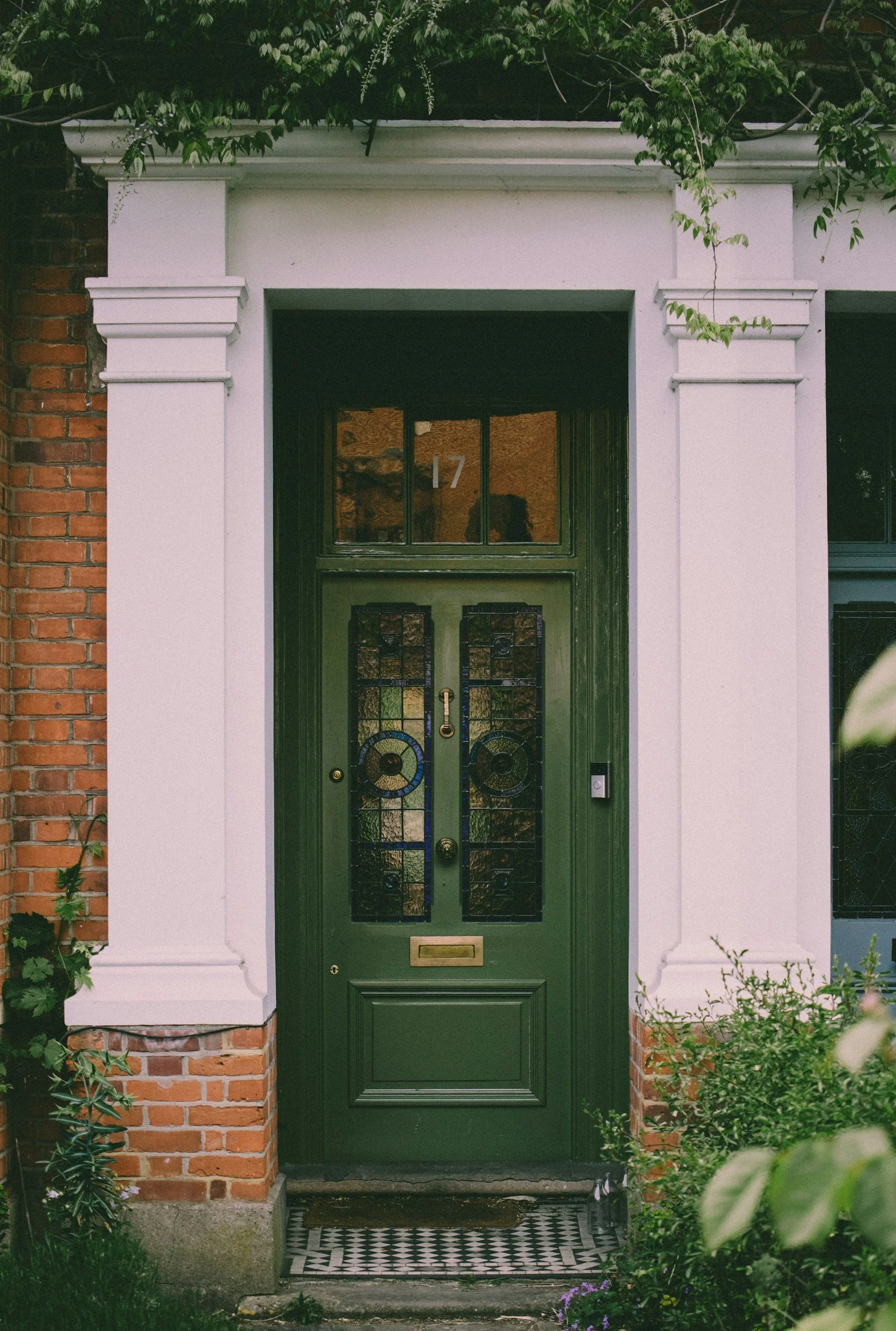 Front entrance of a house with a green door, decorative stained glass panels, and a gold mailbox. The house has a brick and white plaster facade with green plants around the doorway.