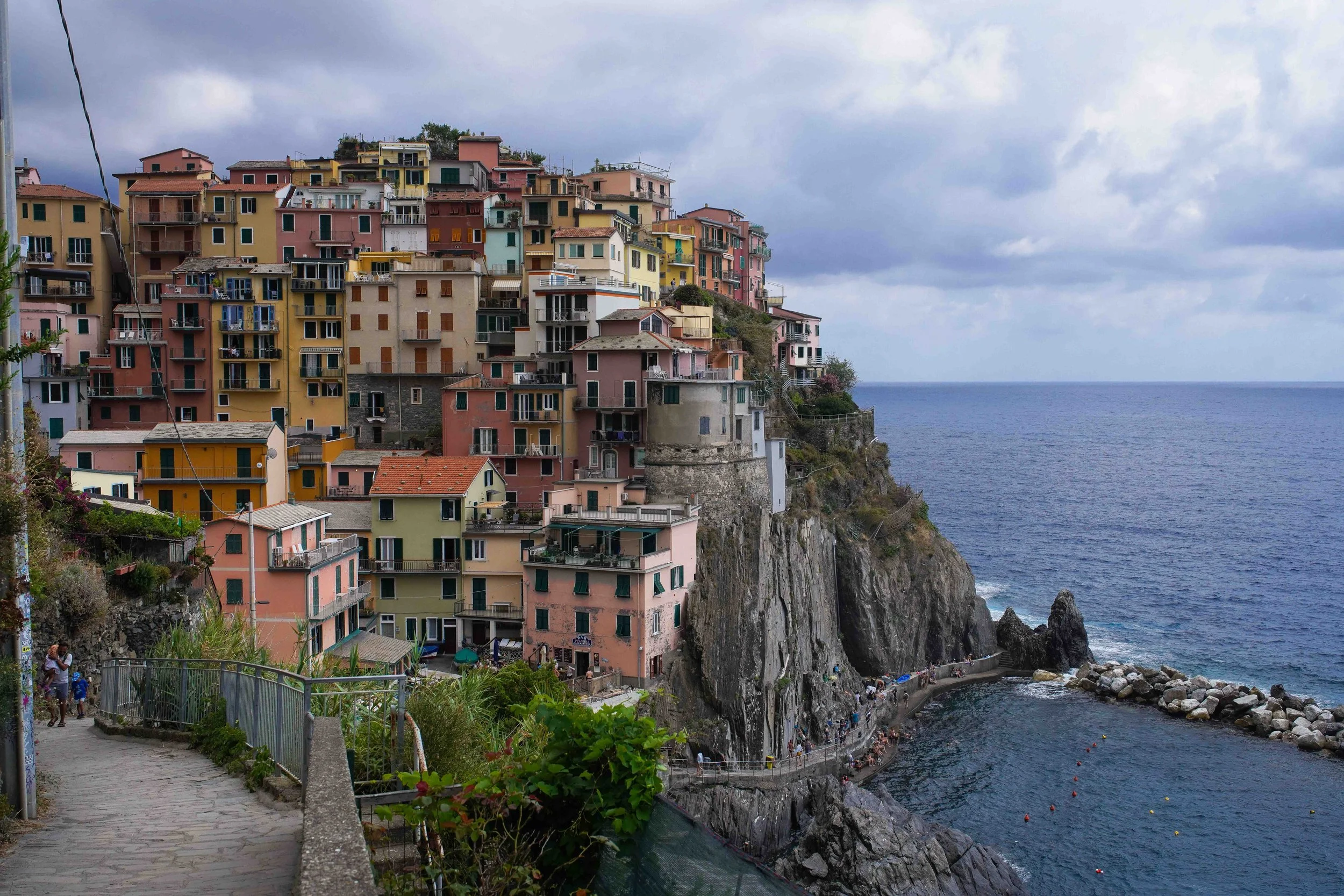 Colorful houses built on a cliffside overlooking the ocean, with a walkway and rocks at the bottom, under a cloudy sky.