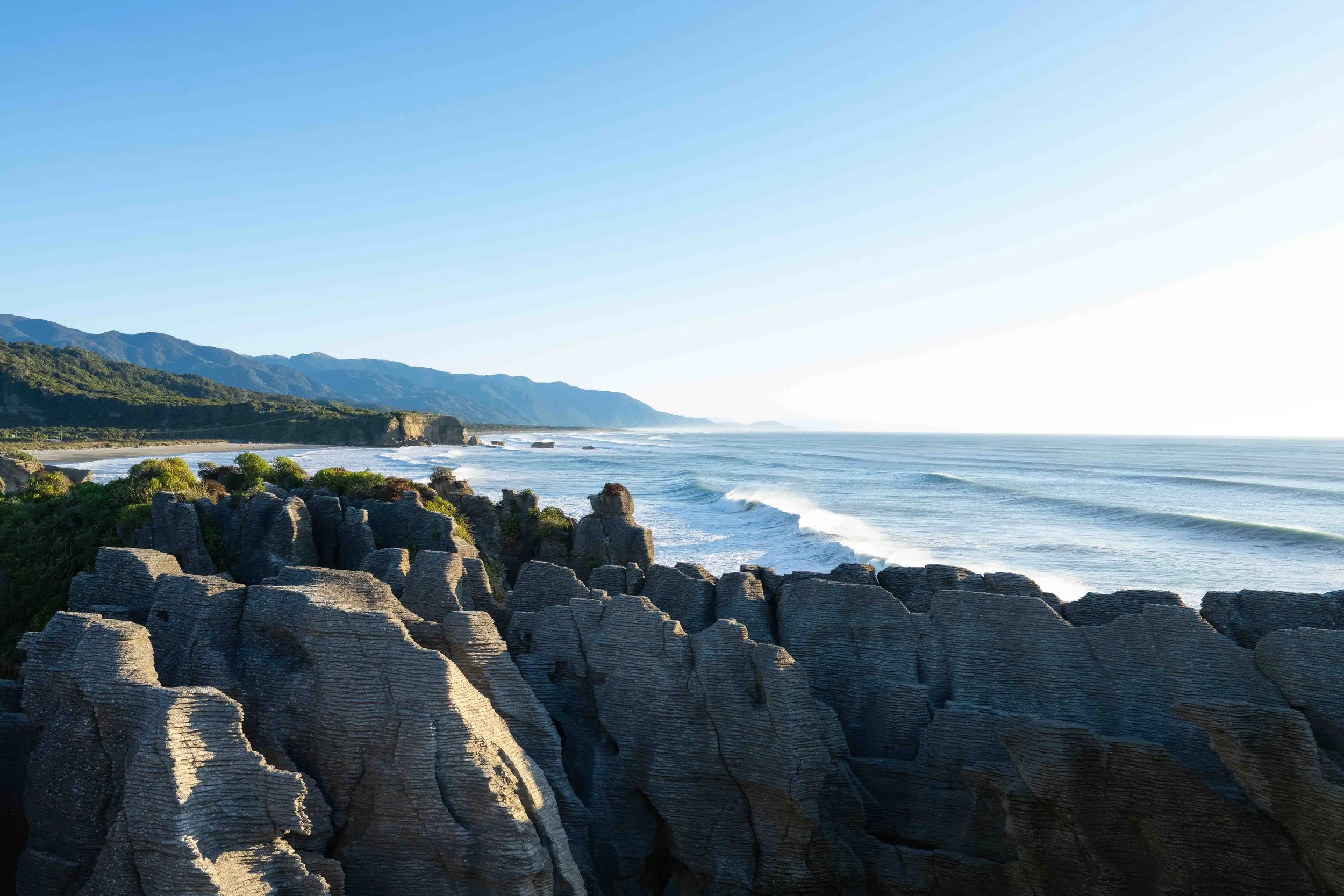 Rock formations along a coastline with the ocean, mountains in the background, and clear skies.