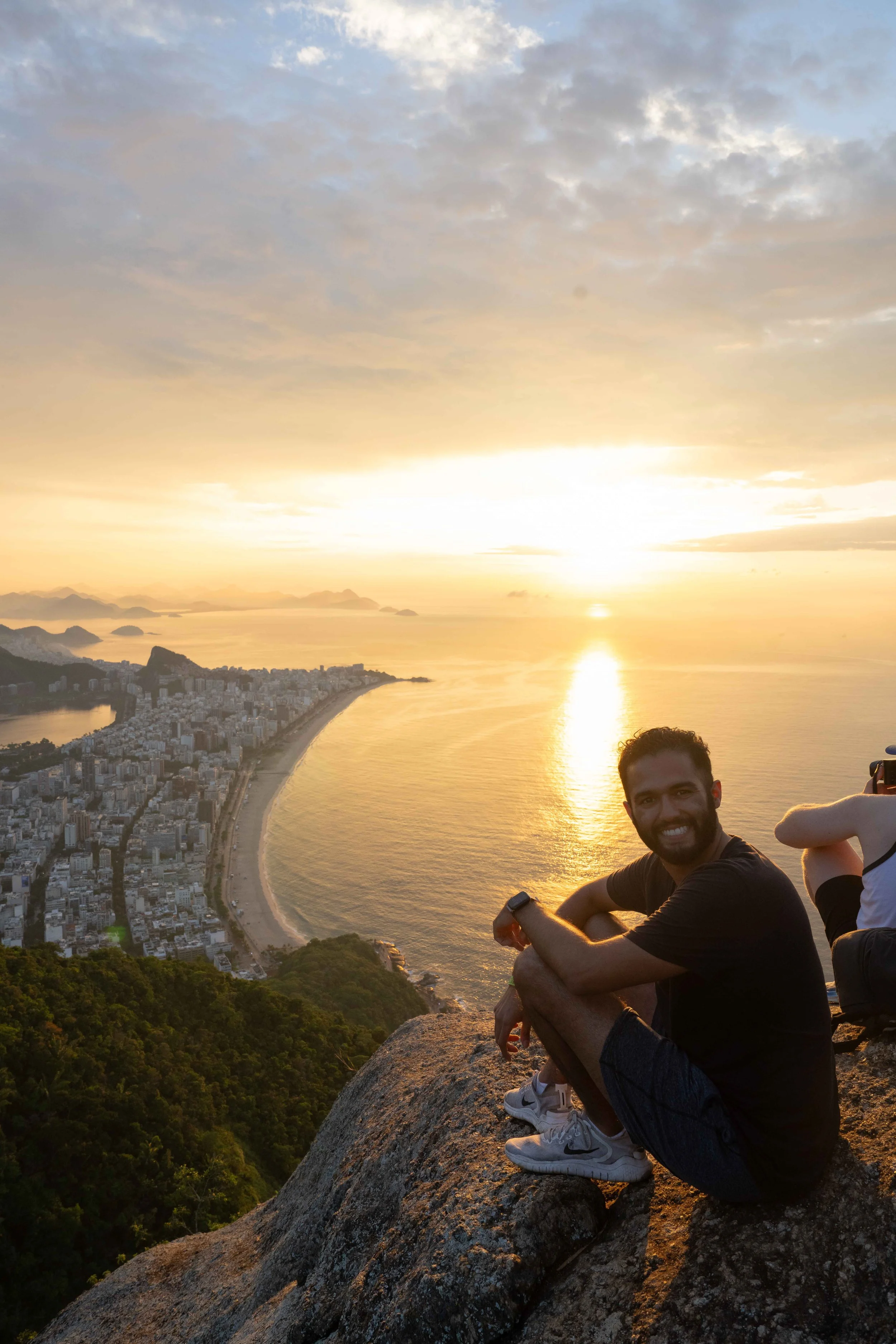 Kunal Sharma sitting on a large rock at a high vantage point in Rio De Janeiro during sunrise, with a view of the coastal city, Ipanema and Copacabana beaches, and ocean in the background.