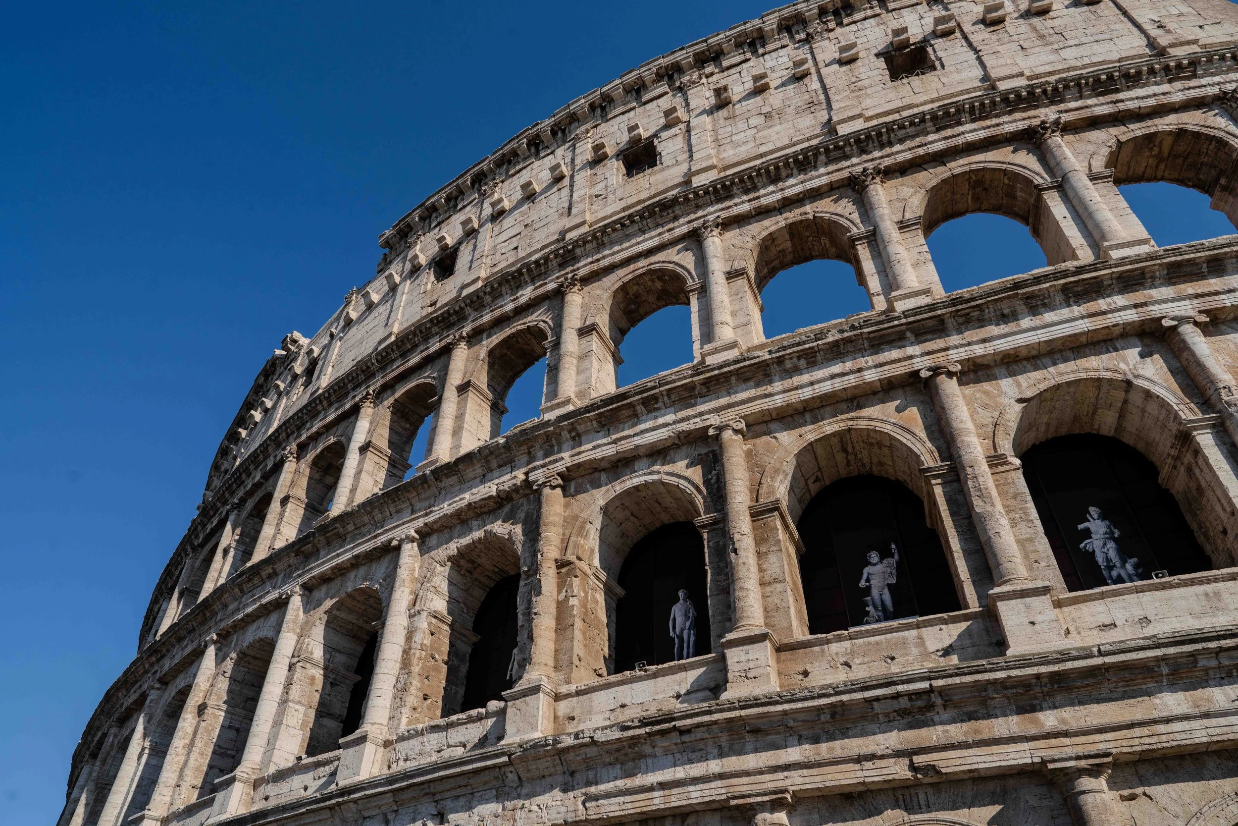 Close-up of the Colosseum's exterior showing arches and statue decorations against a clear blue sky.