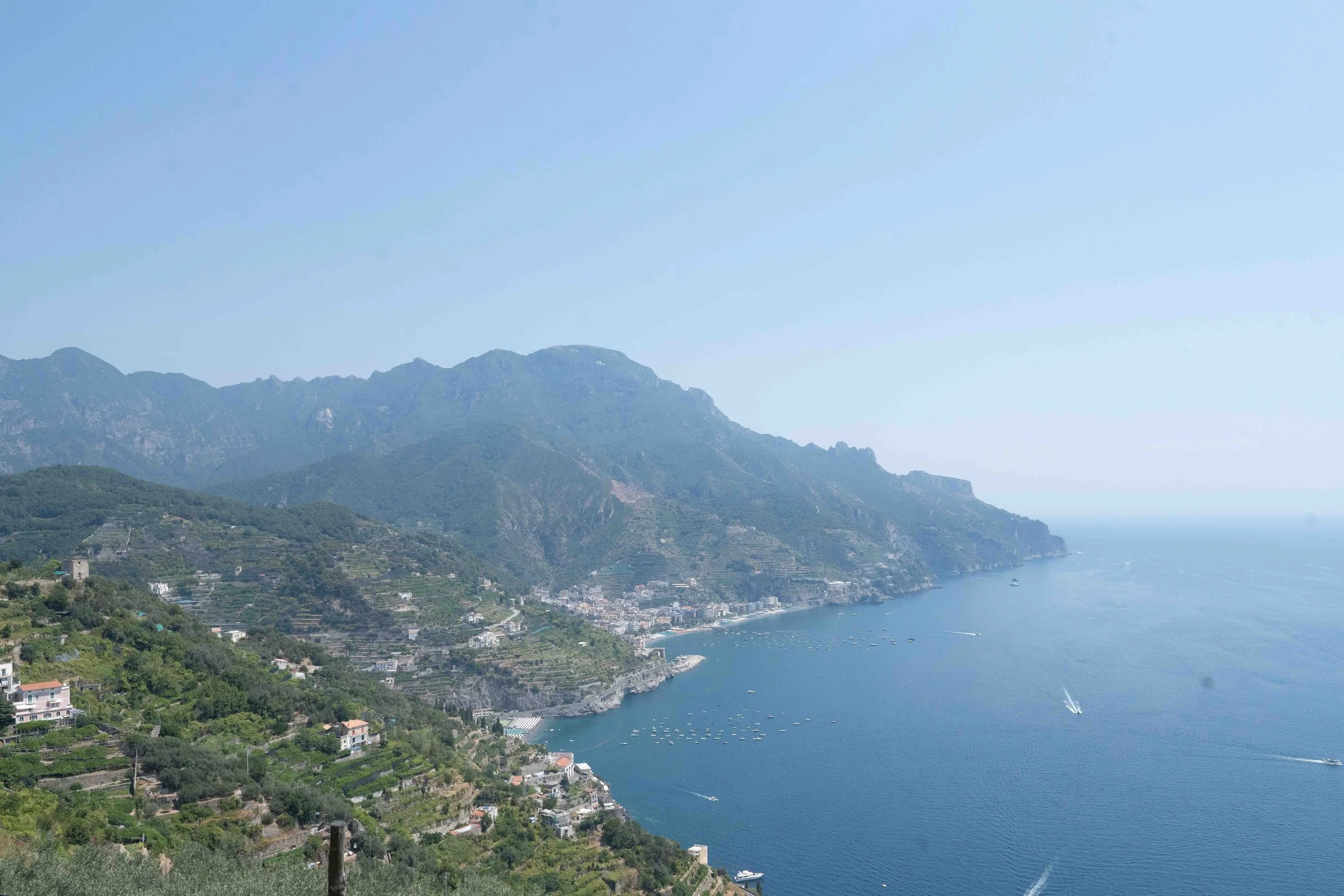 Scenic view of a coastline with green hills and residential buildings, a mountain range in the background, and boats in the water under a clear blue sky.