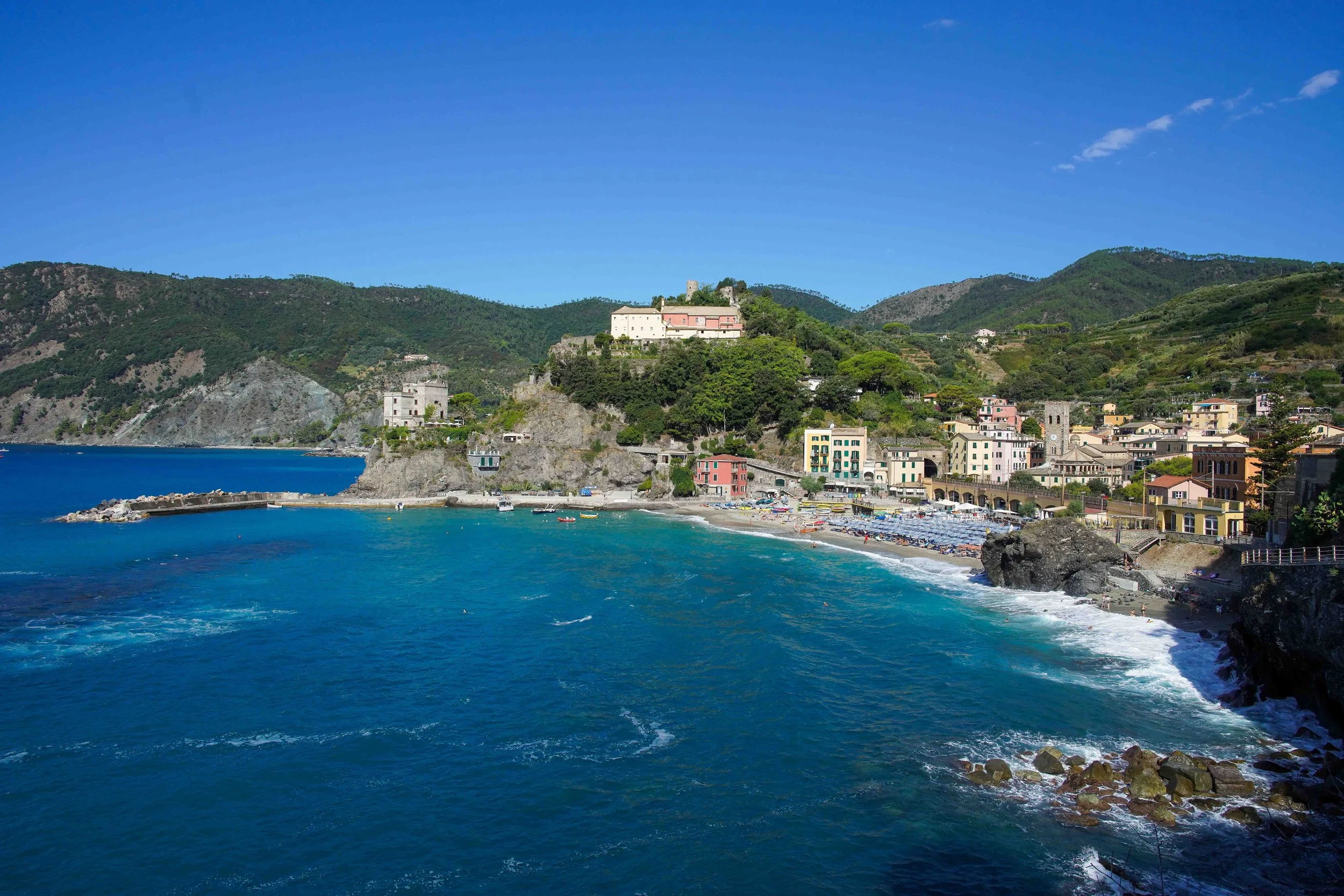 Monterosso al Mare, Cinque Terre, Italy, with colorful buildings along a shoreline, with green hills and mountains in the background under a clear blue sky.