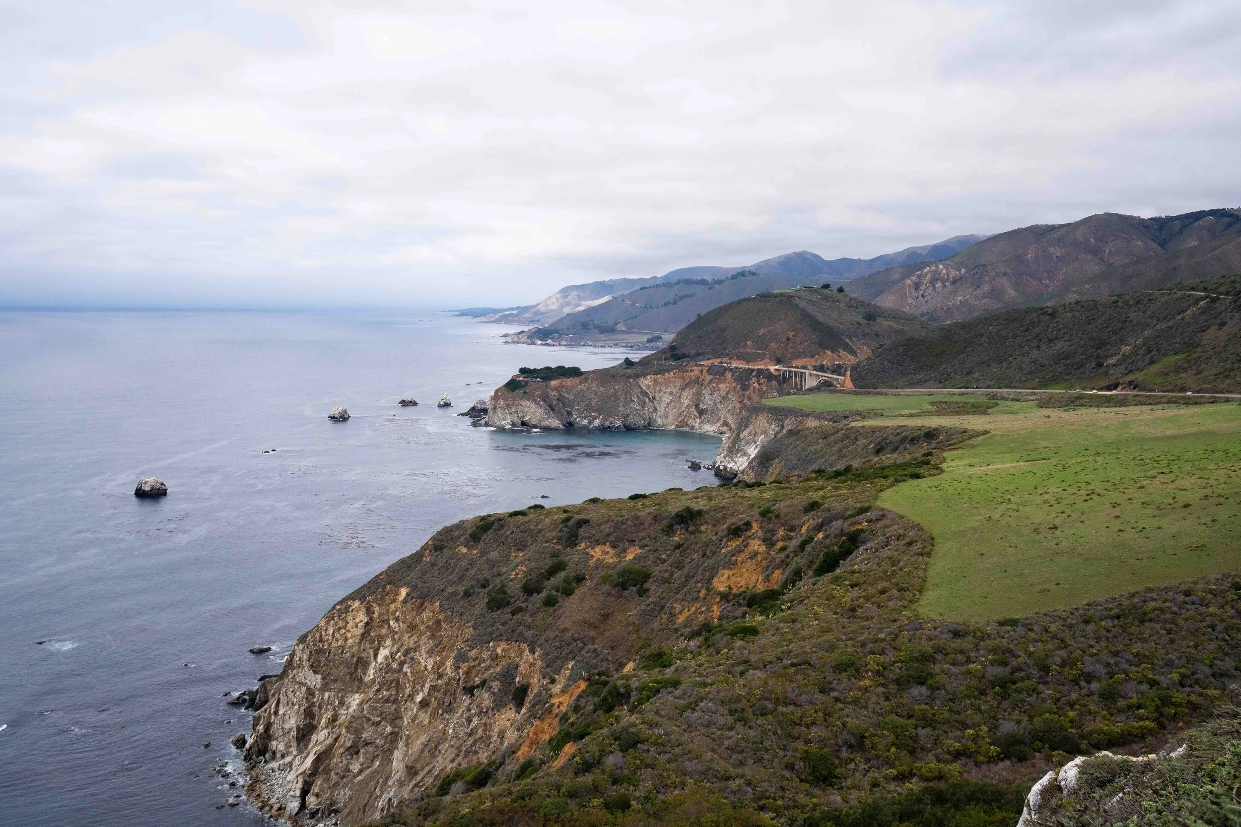 A coastal landscape with cliffs, greenery, ocean waves, and distant mountains under a cloudy sky.