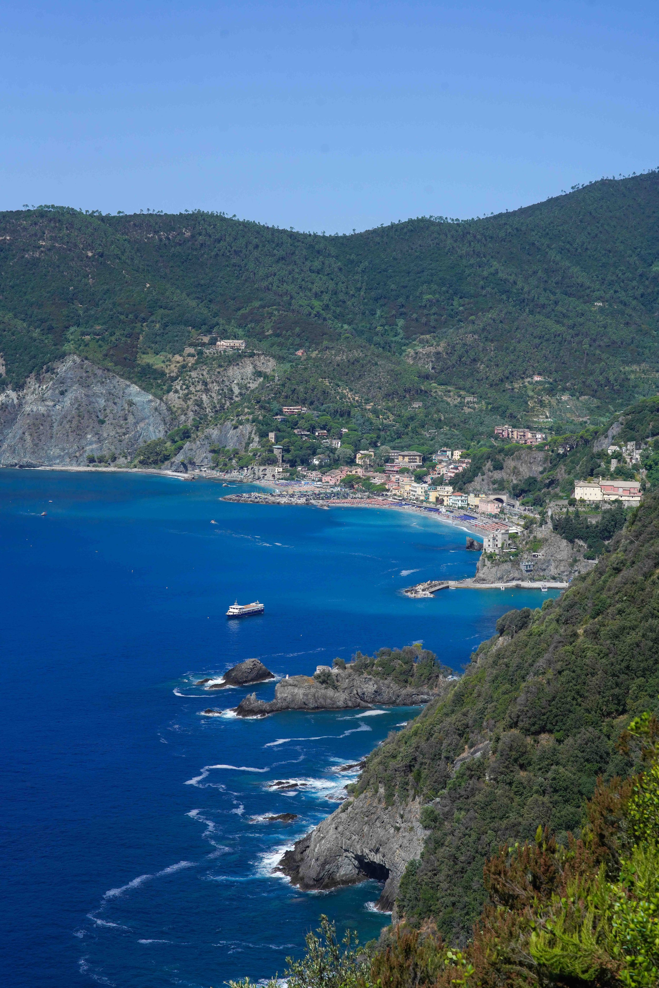 Monterosso al Mare, Cinque Terre, Italy, with a deep blue sea, rocky shoreline, green hills, and a boat is sailing in the water.