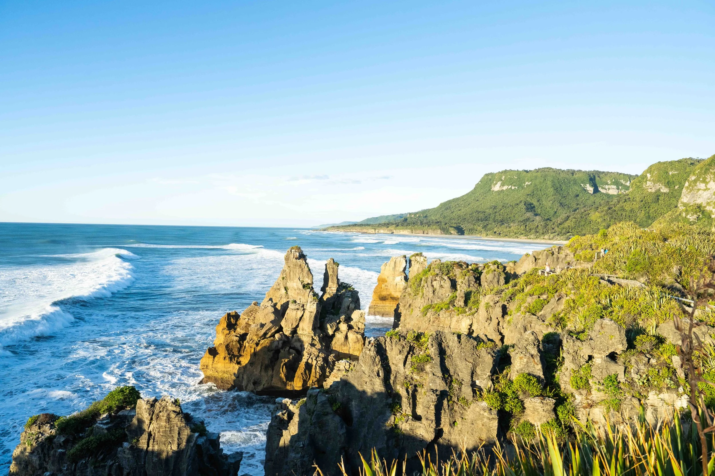 Coastal landscape with rocky cliffs, green hills in the distance, and ocean waves crashing against the rocks, under a clear blue sky.