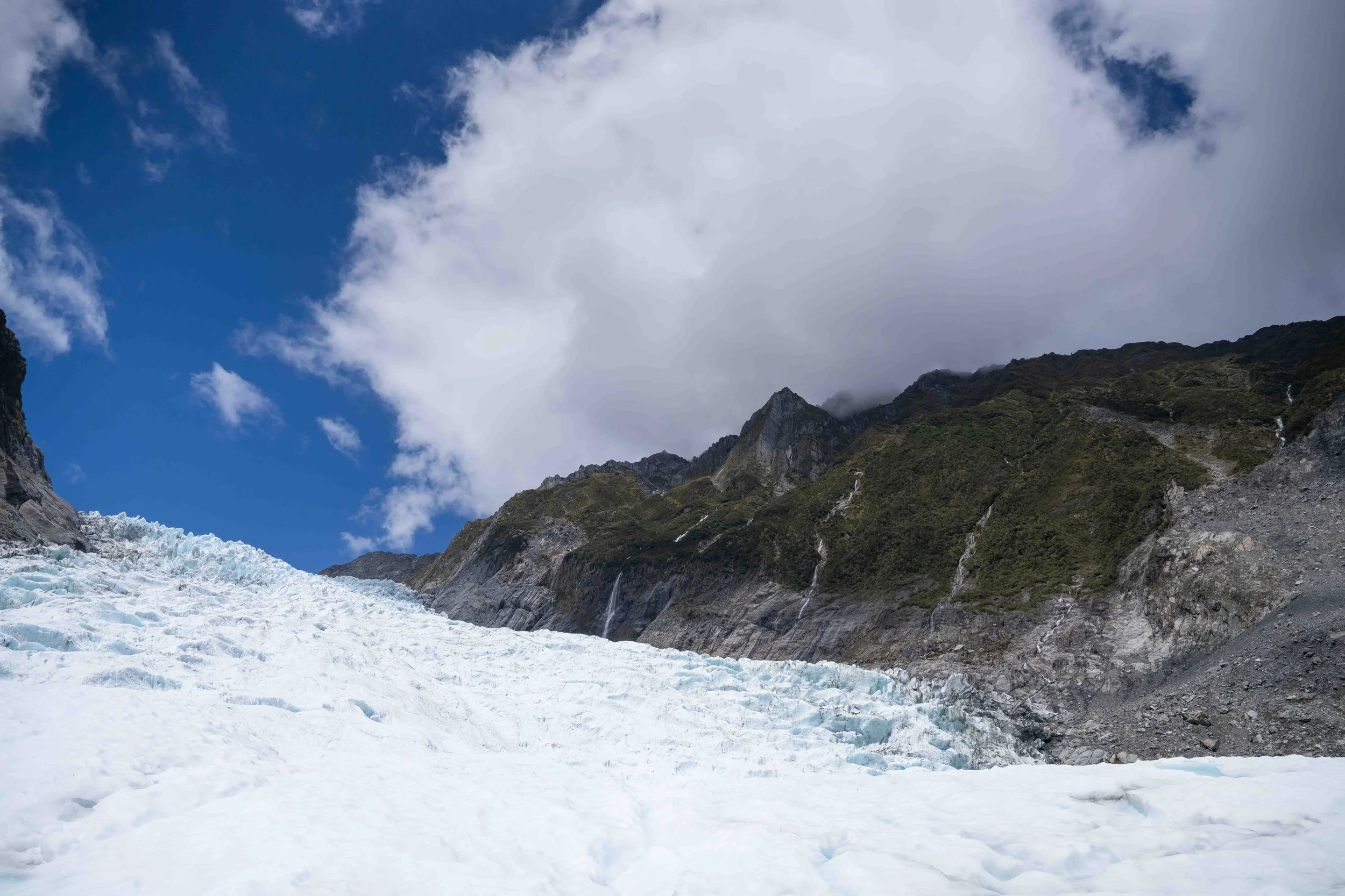 A glacier with ice and snow at the base of rugged green mountains under a partly cloudy sky.