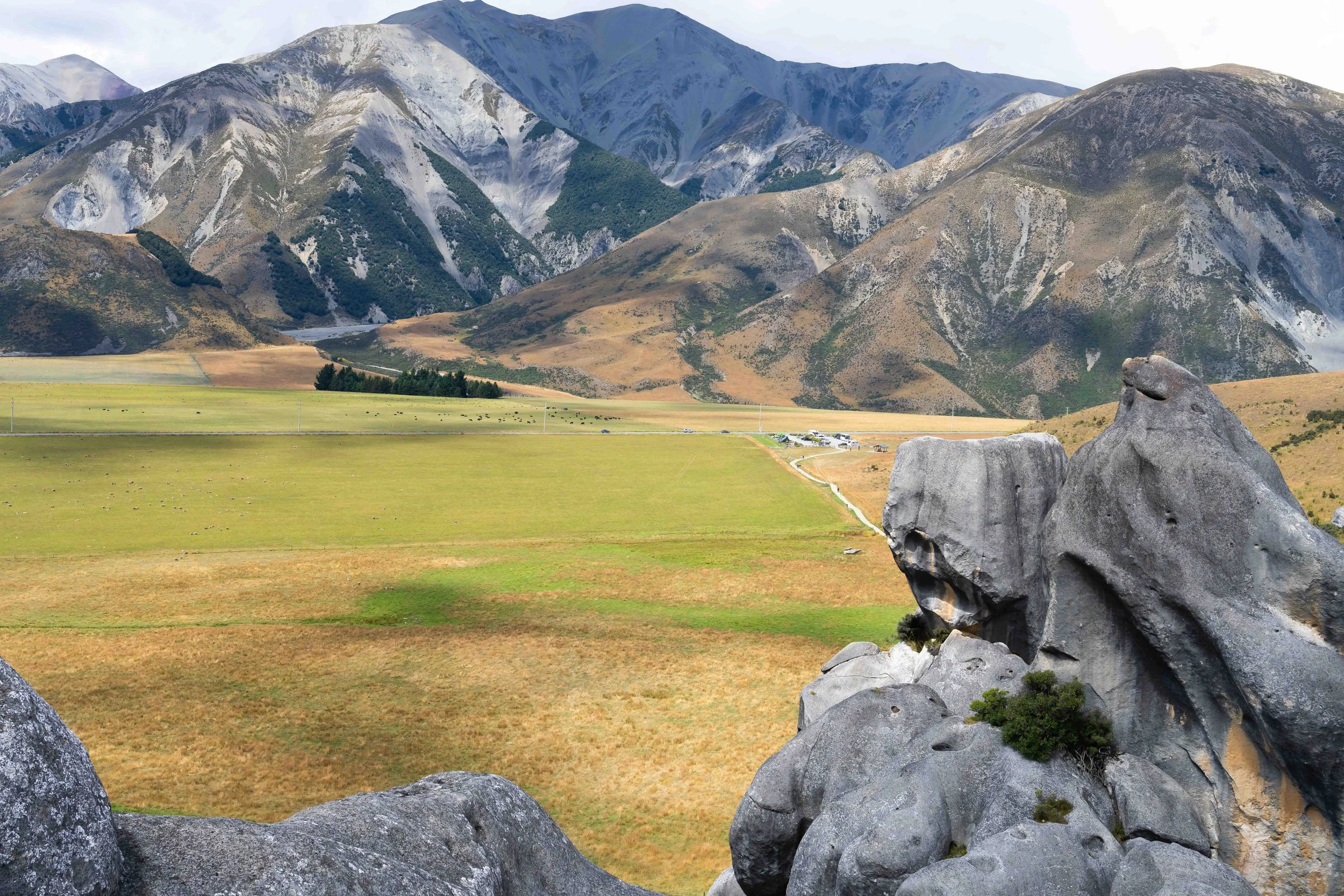 View of mountains from Castle Rock, New Zealand, with rocky slopes and green patches, a valley with grassy fields, and large rocks in the foreground.