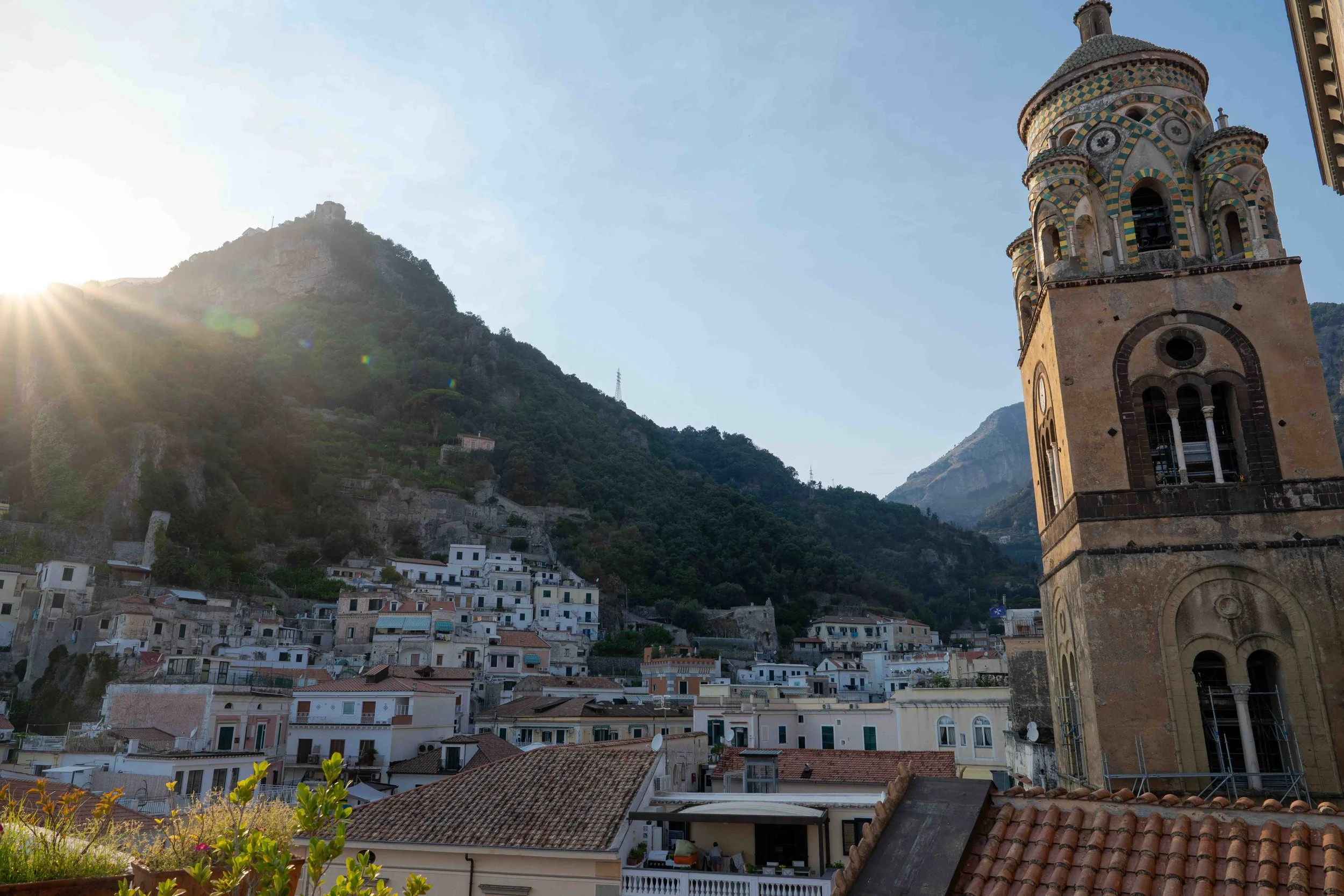 Scenic view of a hillside town with a tall historic church tower in the foreground, with colorful houses, lush greenery, and a mountain backdrop under a bright blue sky with the sun shining.