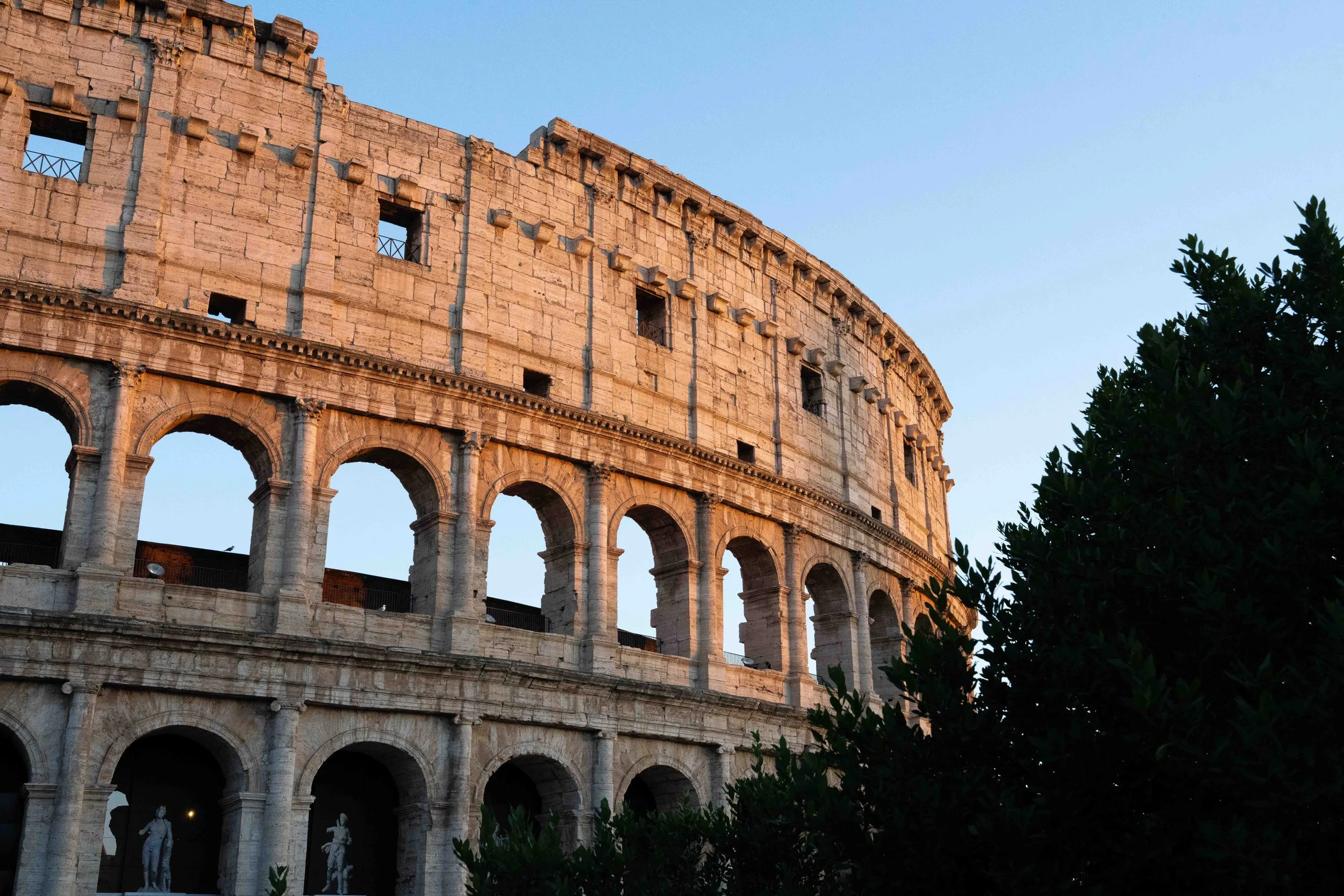 The image shows the exterior of the Roman Colosseum at sunset, with parts of the ancient stone structure highlighted by sunlight. Green trees are partially visible on the right side of the photo, and the sky is clear and blue.