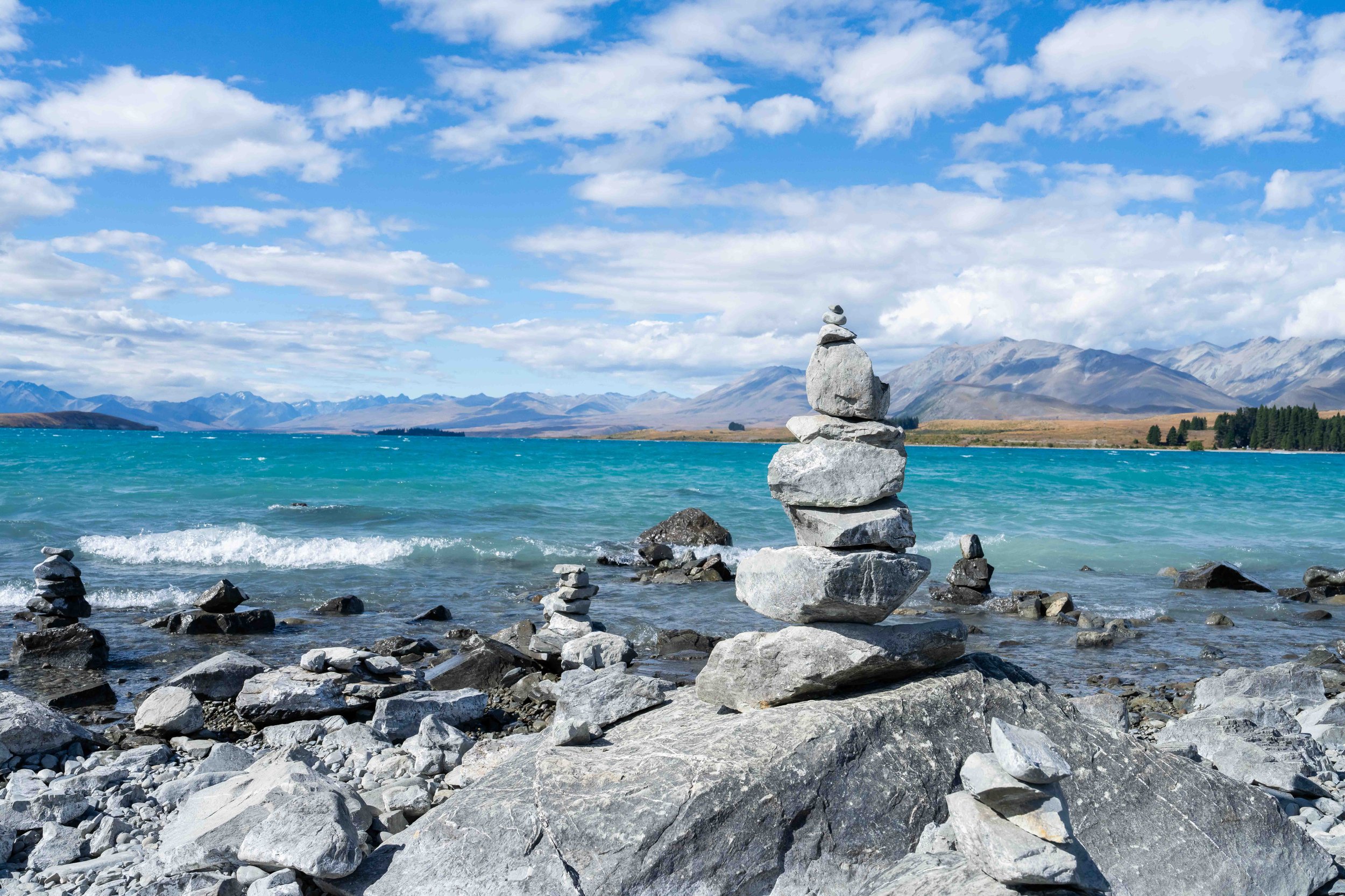 Stacked rocks on the shore at Lake Tekapo, New Zealand, with mountains in the background and a partly cloudy sky.