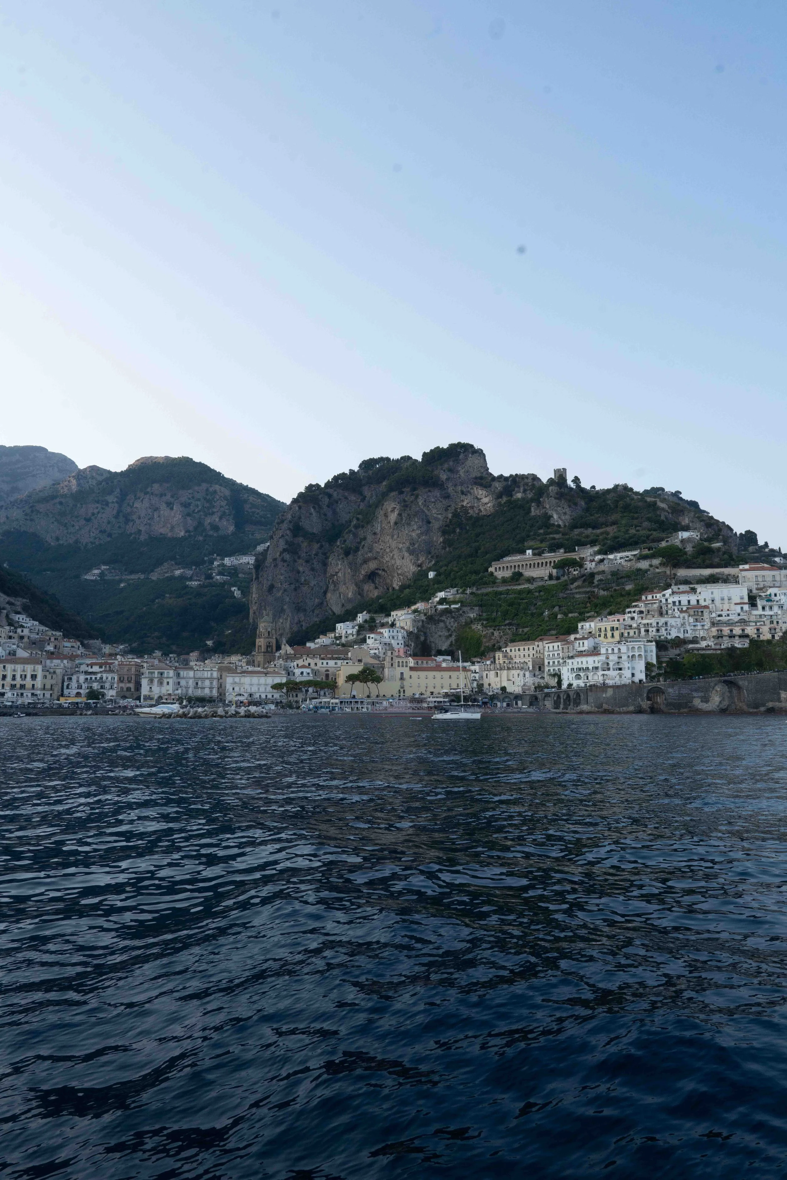 View of a Amalfi, Italy with colorful buildings on a hillside, rocky cliffs, a tower, and boats on dark water under a clear blue sky.