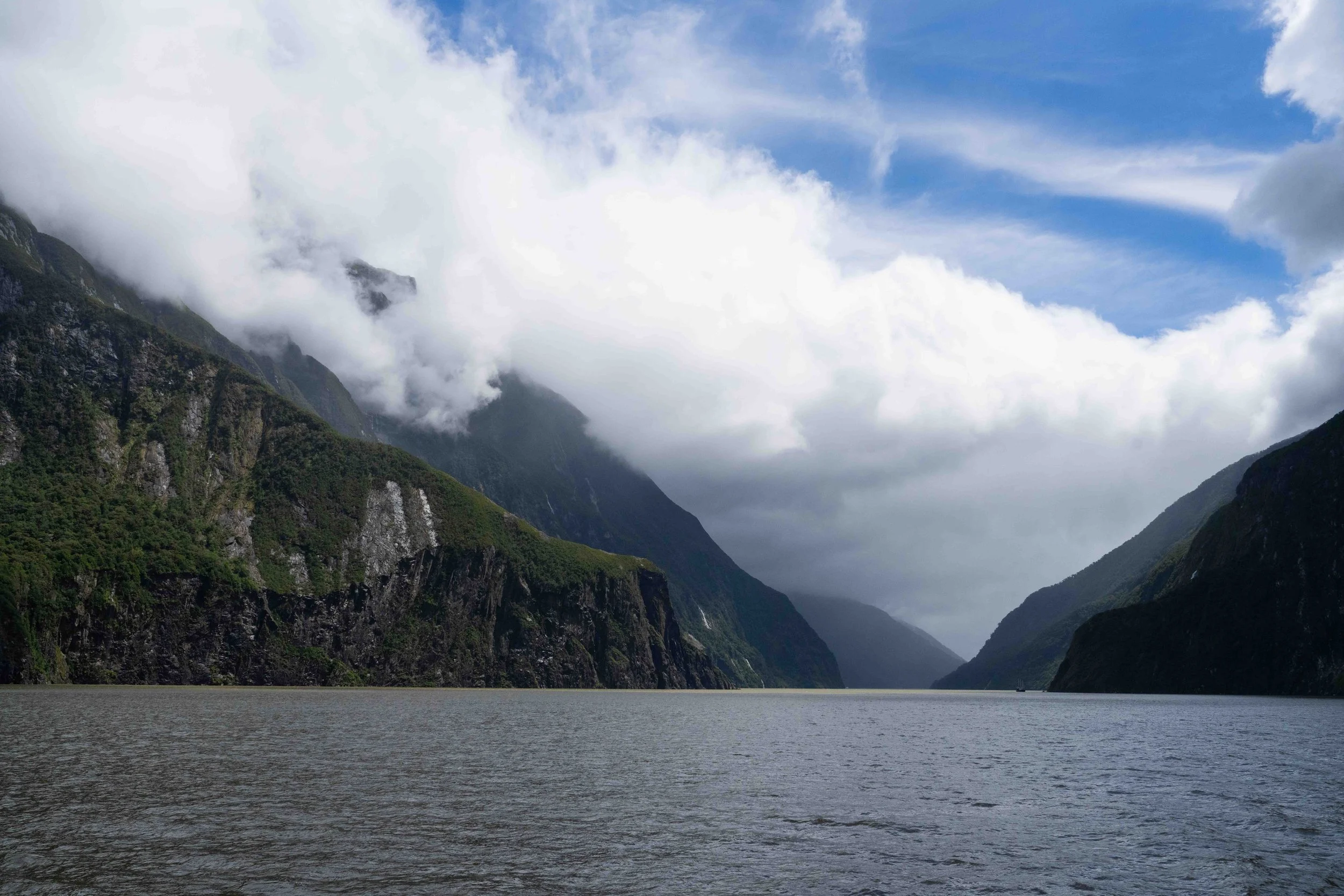 Scenic view of a river flowing between lush green mountains with clouds covering the peaks and a partly cloudy blue sky.