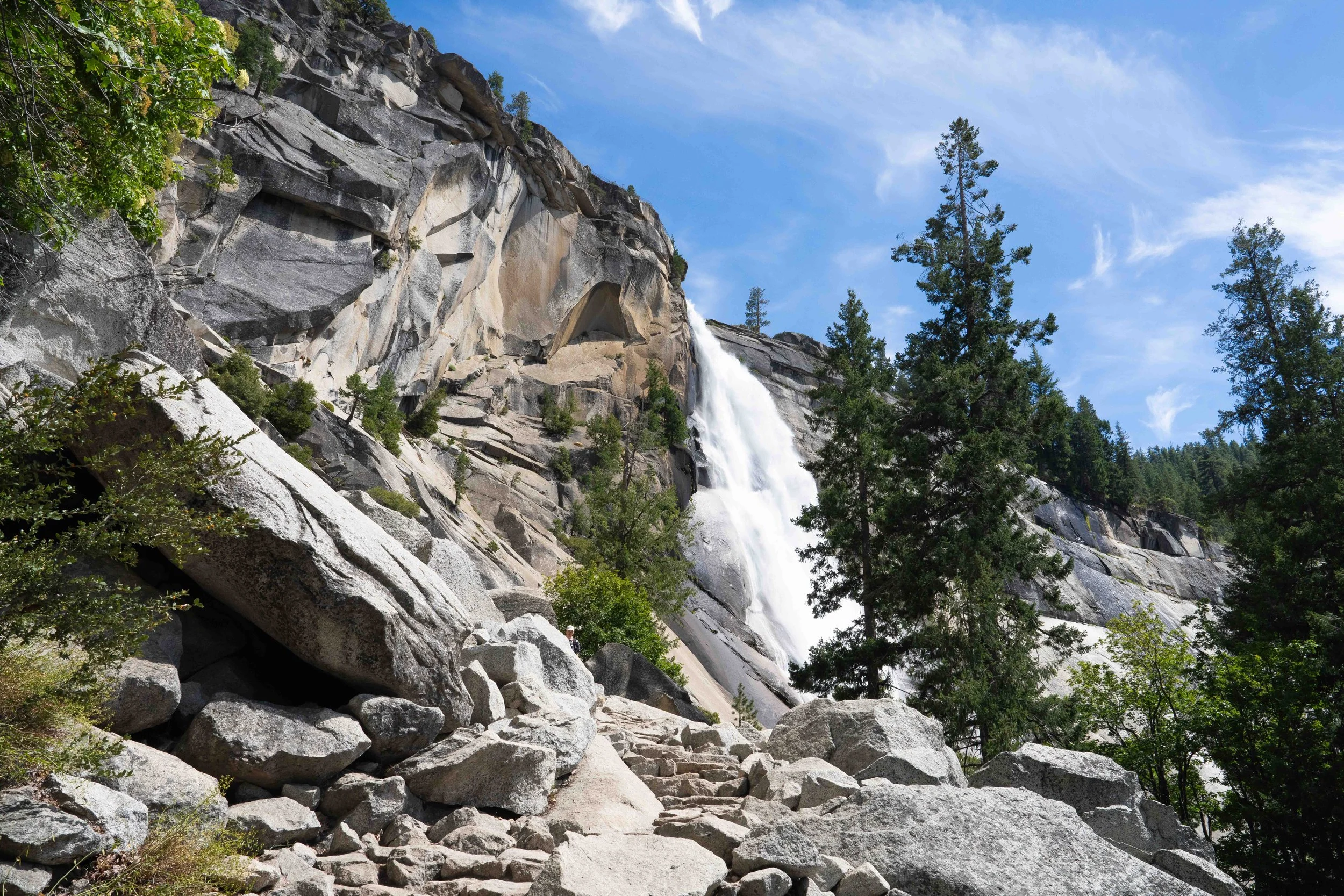 A scenic landscape of granite rocks and boulders with a waterfall flowing down a steep cliff surrounded by a forest of tall pine trees, under a partly cloudy blue sky.