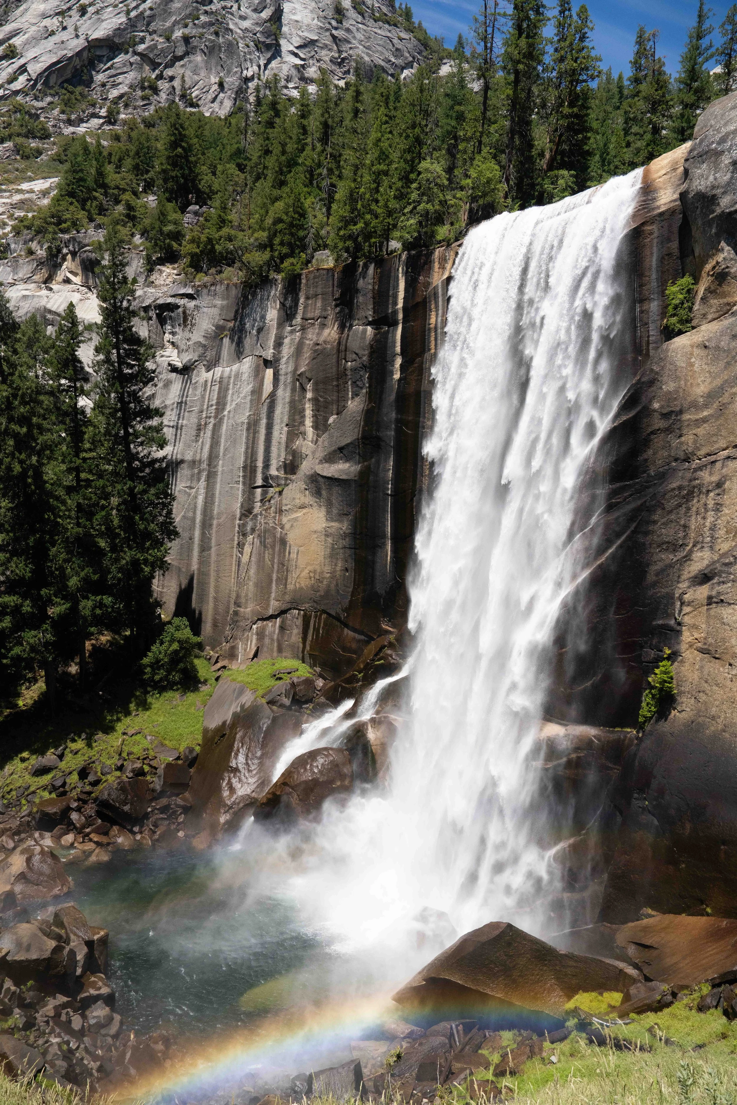 A tall waterfall cascading down a rocky cliff with greenery and trees around it, creating a rainbow at the base of the waterfall.
