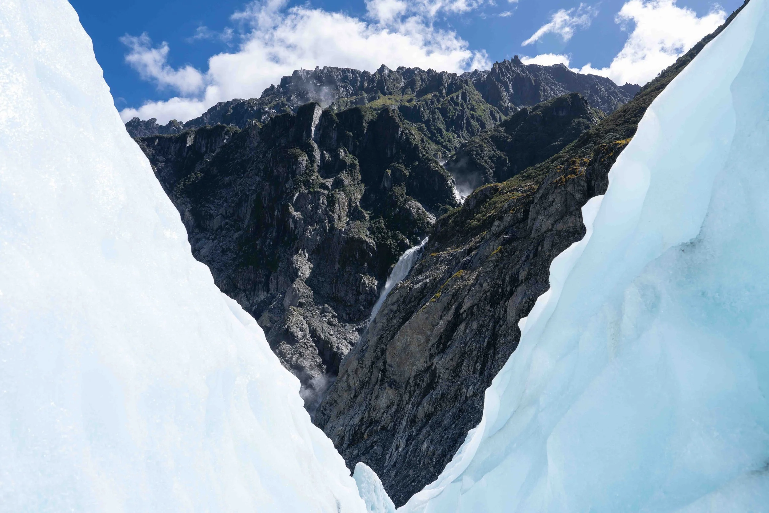 A scenic mountain landscape viewed through a break in large ice formations, showing rugged green mountains with rocky peaks under a partly cloudy sky.