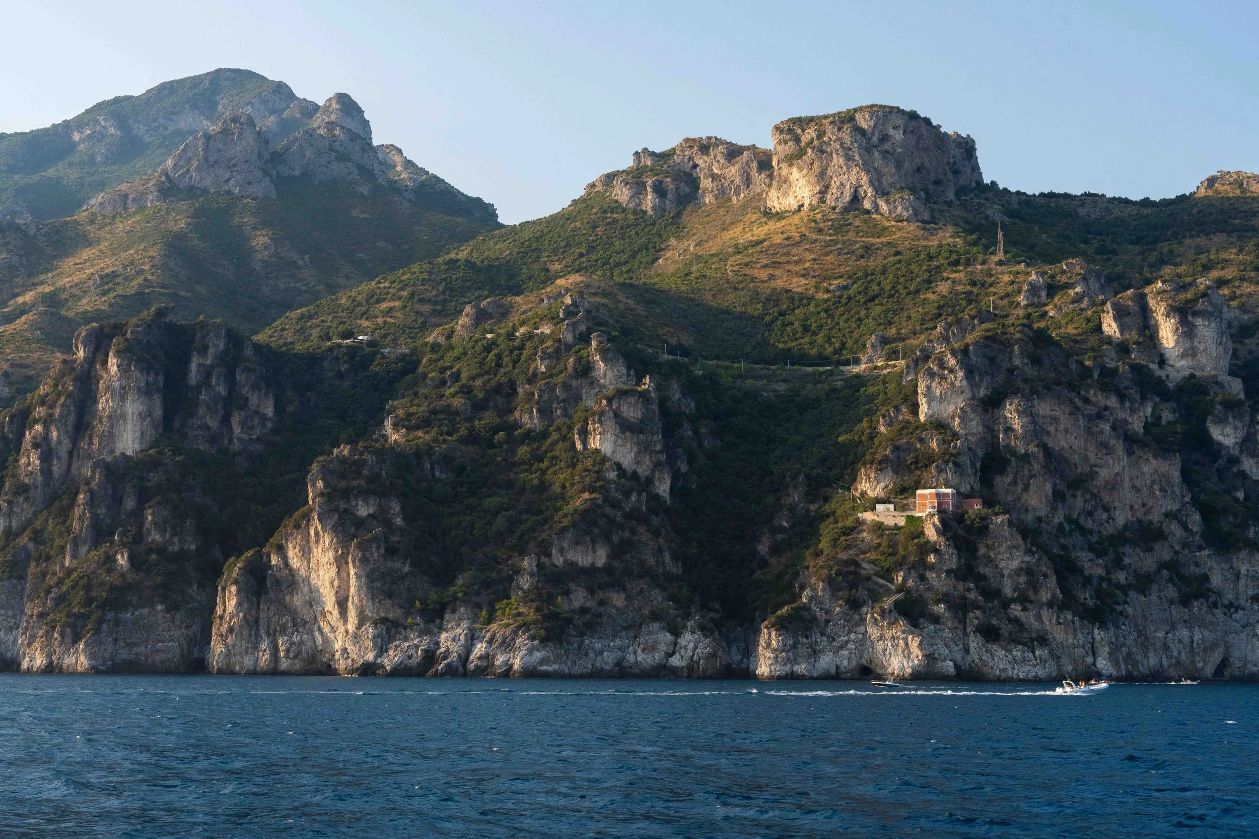 A scenic view of rugged green mountains in Italy's Amalfi Coast, rising sharply from the sea, with a small building on one of the lower slopes and a boat speeding across the water in the foreground.