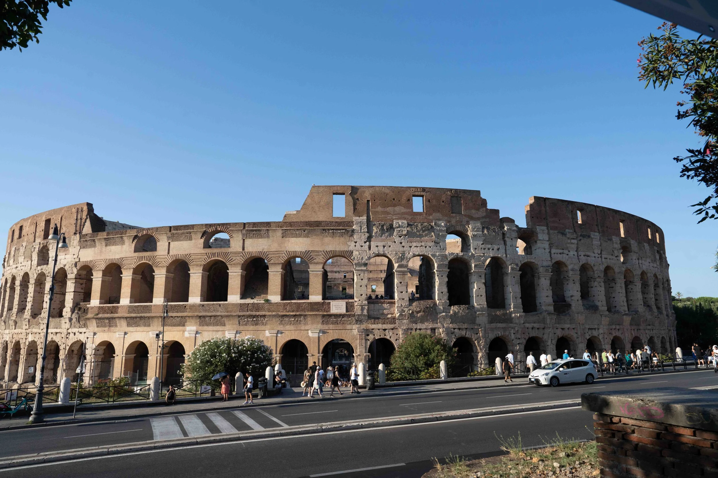 The image shows the Roman Colosseum, an ancient amphitheater in Rome, Italy, under a clear blue sky, with tourists walking along the street in front.
