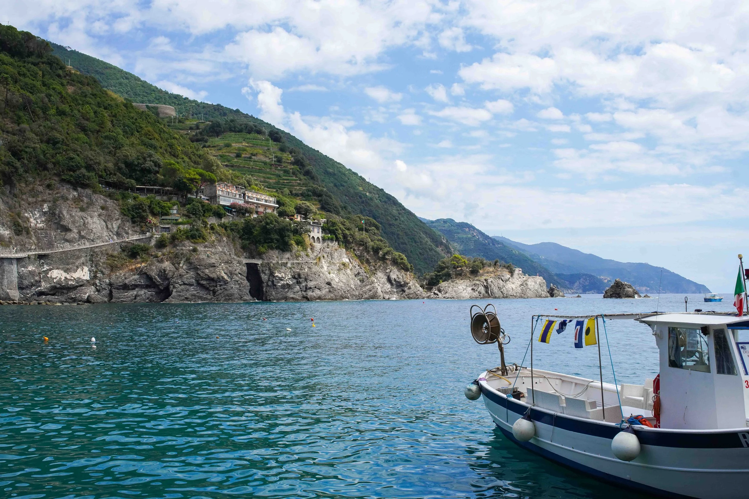 View of a rocky coastline in Monterosso al Mare, Cinque Terre, Italy, with green hillside and houses, small boats on calm water, and a cloudy sky.