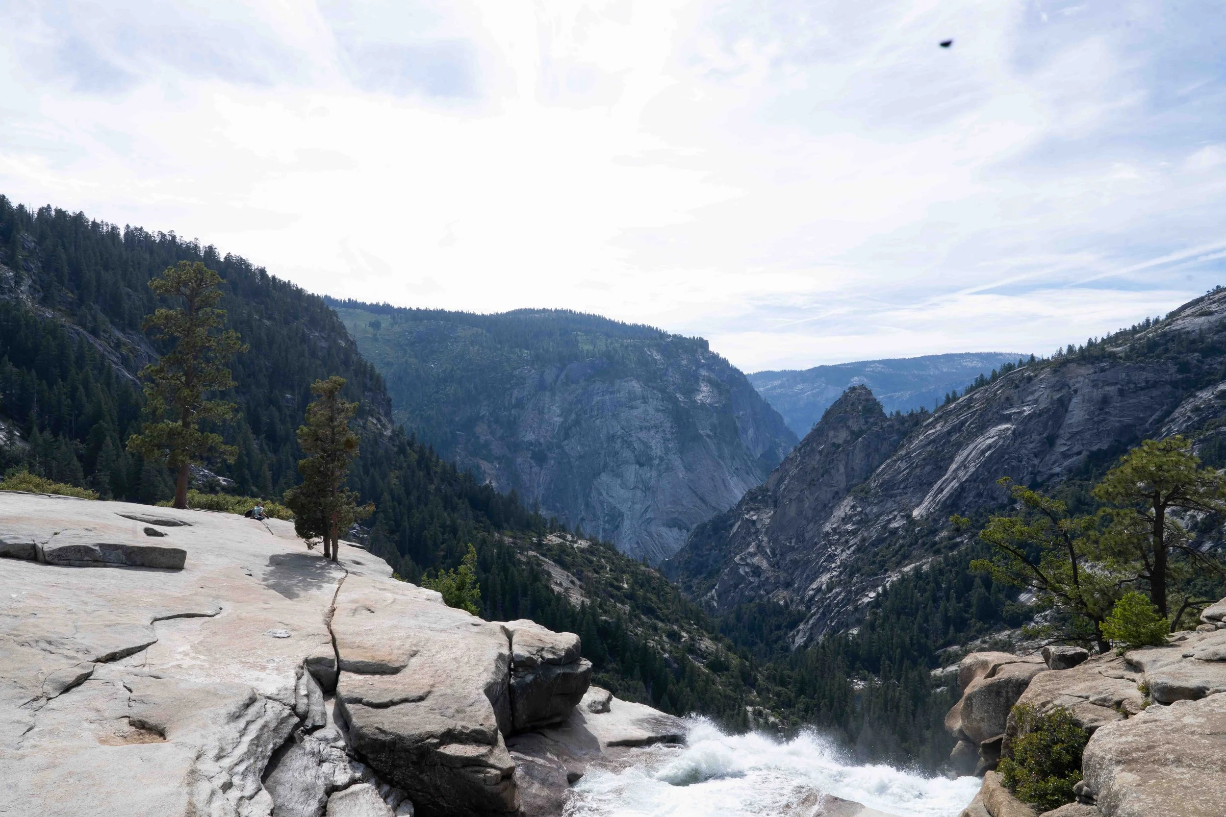 Scenic mountain landscape with towering cliffs, dense pine forests, and a rushing river through a rocky valley, under a partly cloudy sky.